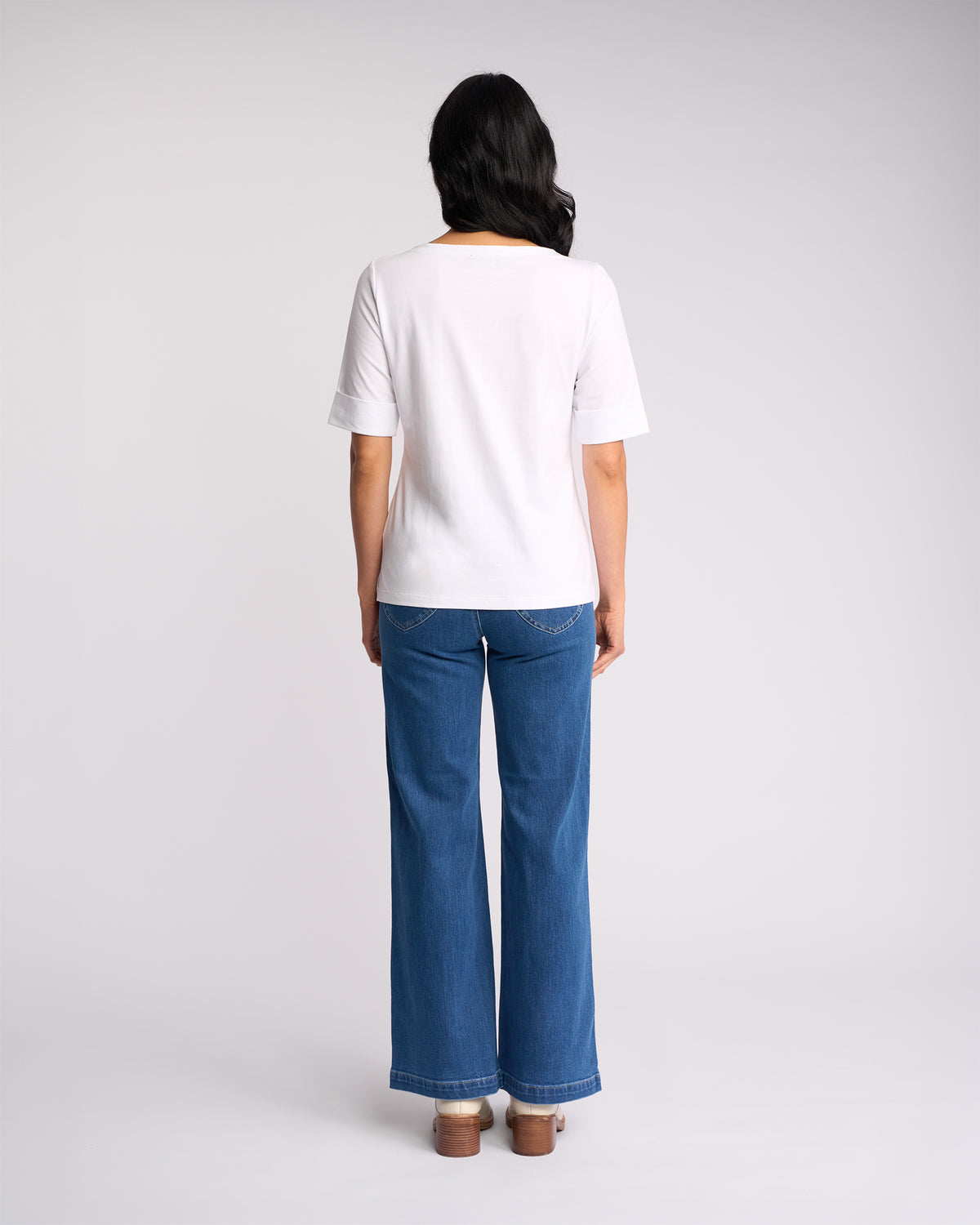 A person with long dark hair stands facing away, wearing the Vassalli Arden Top in white, paired with blue wide-leg jeans and brown shoes, against a plain light-colored background.