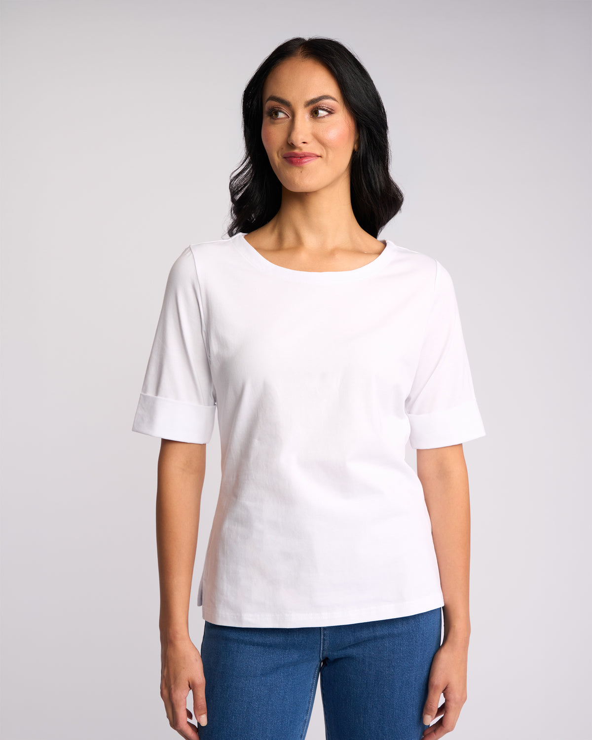 A woman with long dark hair wears the Vassalli Arden Top - White and blue jeans, standing against a plain light background, looking slightly to her left with a gentle smile.