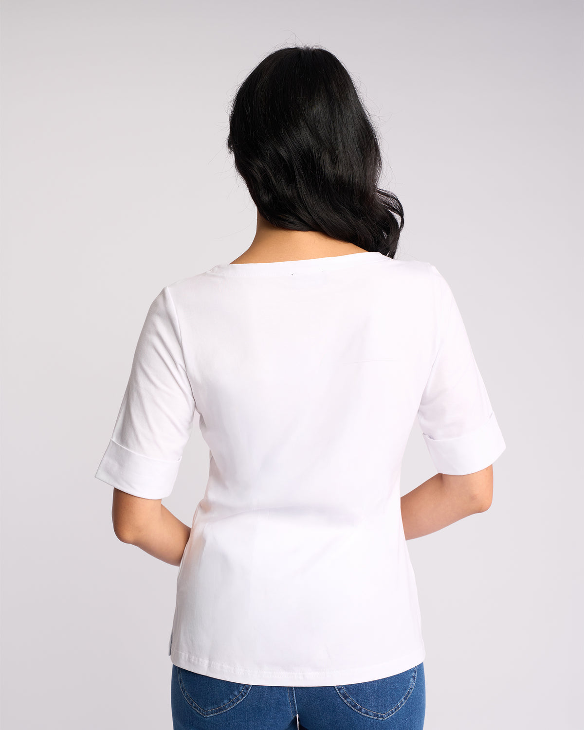 A woman with long dark hair stands with her back to the camera, wearing a Vassalli Arden Top - White and blue jeans, against a light gray background.
