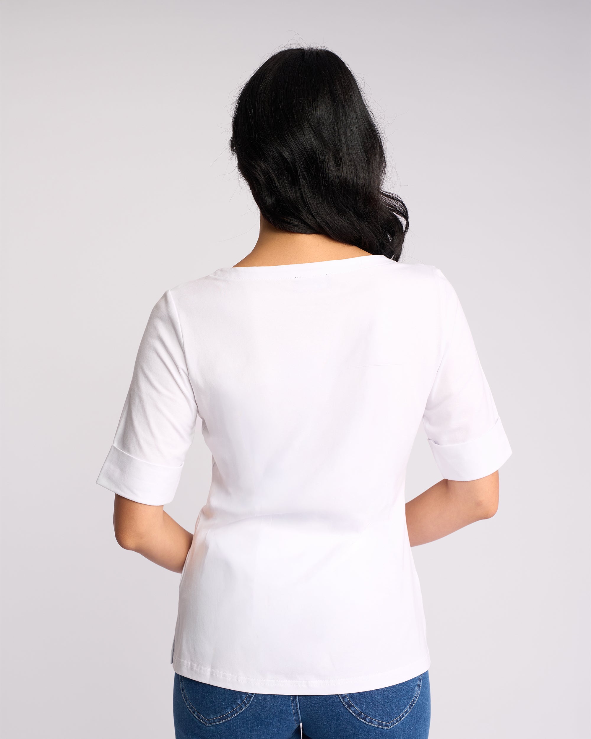A woman with long dark hair stands with her back to the camera, wearing a Vassalli Arden Top - White and blue jeans, against a light gray background.