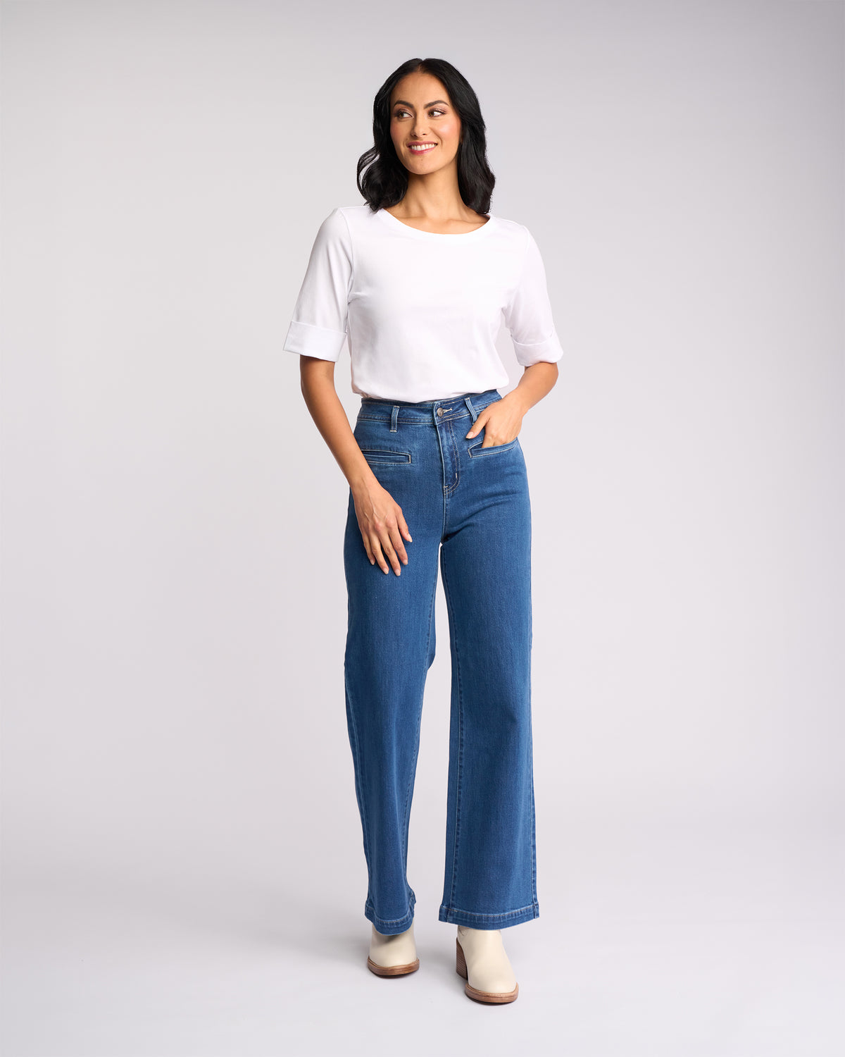 A woman models the Vassalli Arden Top - White, styled with high-waisted blue jeans and light boots. She stands against a plain background, smiling and looking slightly to the side, with long dark hair.