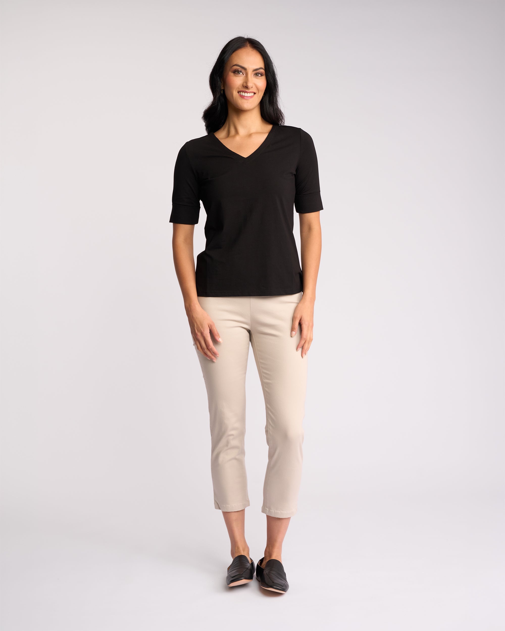 A woman with long dark hair stands smiling, wearing the Vassalli Reece Top in black with light beige cropped pants and black shoes, against a plain light background.