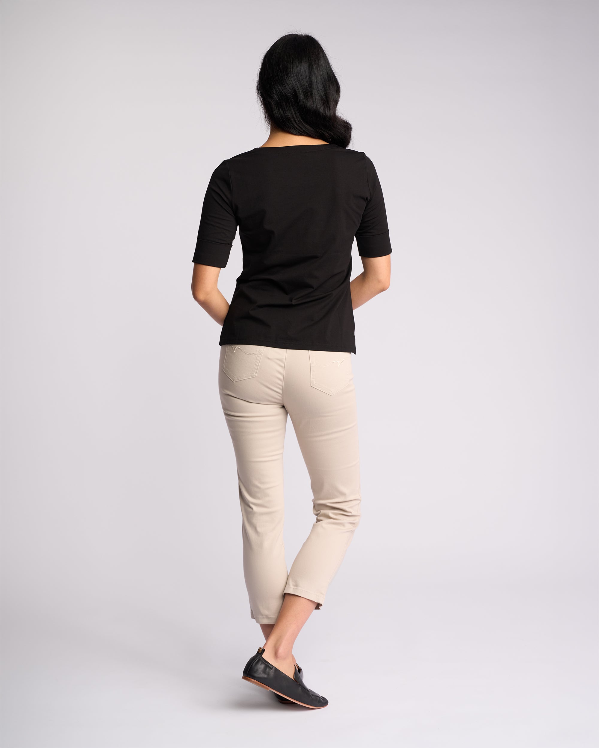 A woman with long dark hair faces away, wearing the Vassalli Reece Top in black—an elegant V-neck style—paired with beige skinny pants and black shoes, showcasing stylish everyday comfort against a plain light background.