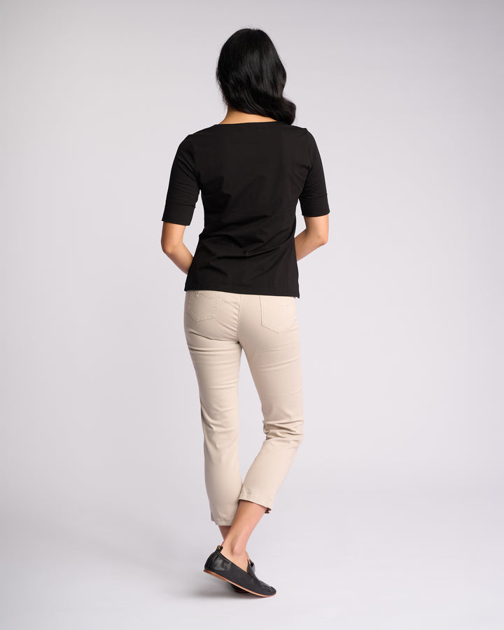 A woman with long dark hair faces away, wearing the Vassalli Reece Top in black—an elegant V-neck style—paired with beige skinny pants and black shoes, showcasing stylish everyday comfort against a plain light background.