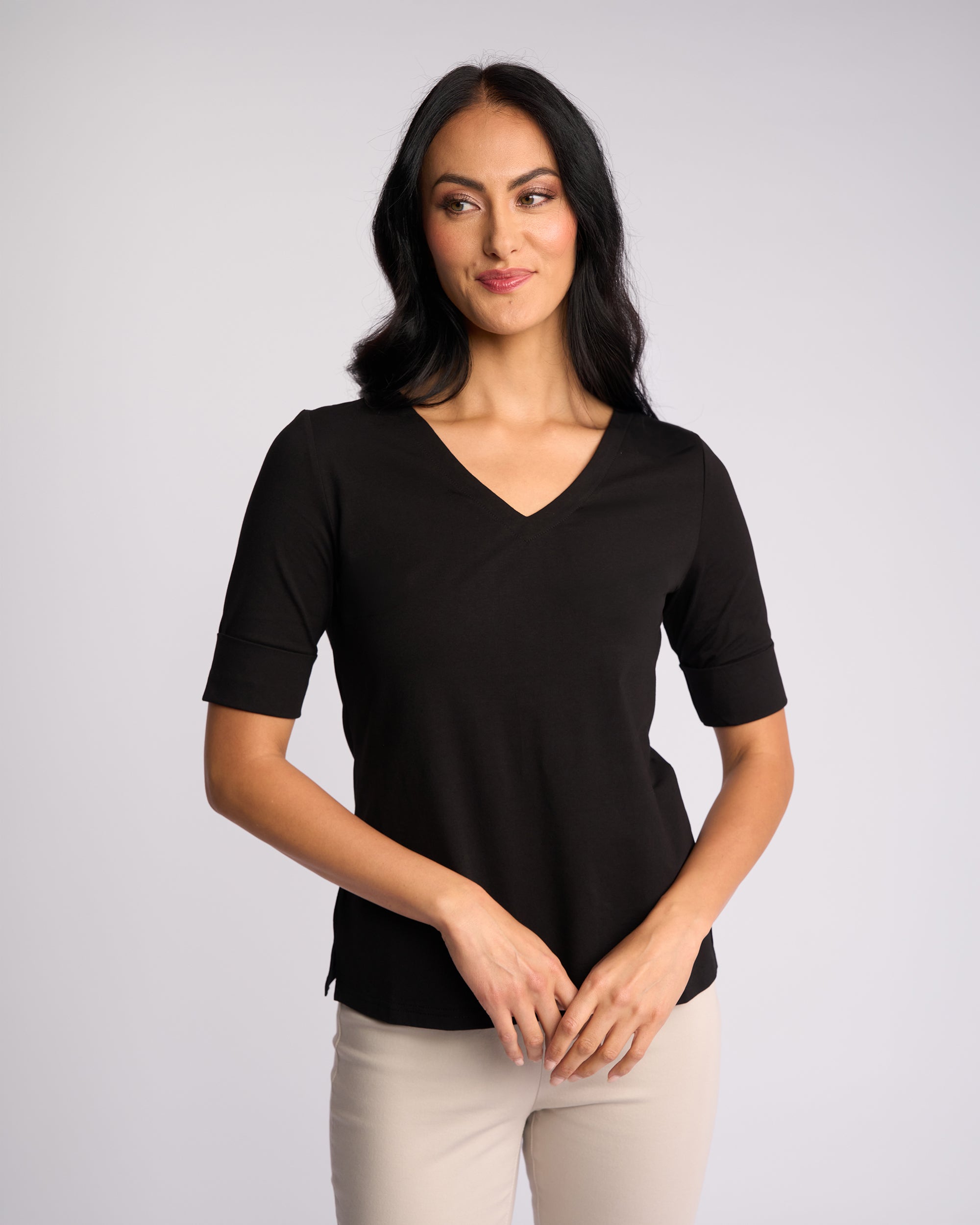 A woman with long dark hair stands against a light background, smiling gently in the Vassalli Reece Top - Black and light pants—her hands loosely clasped, showcasing a relaxed yet stylish look for everyday wear.