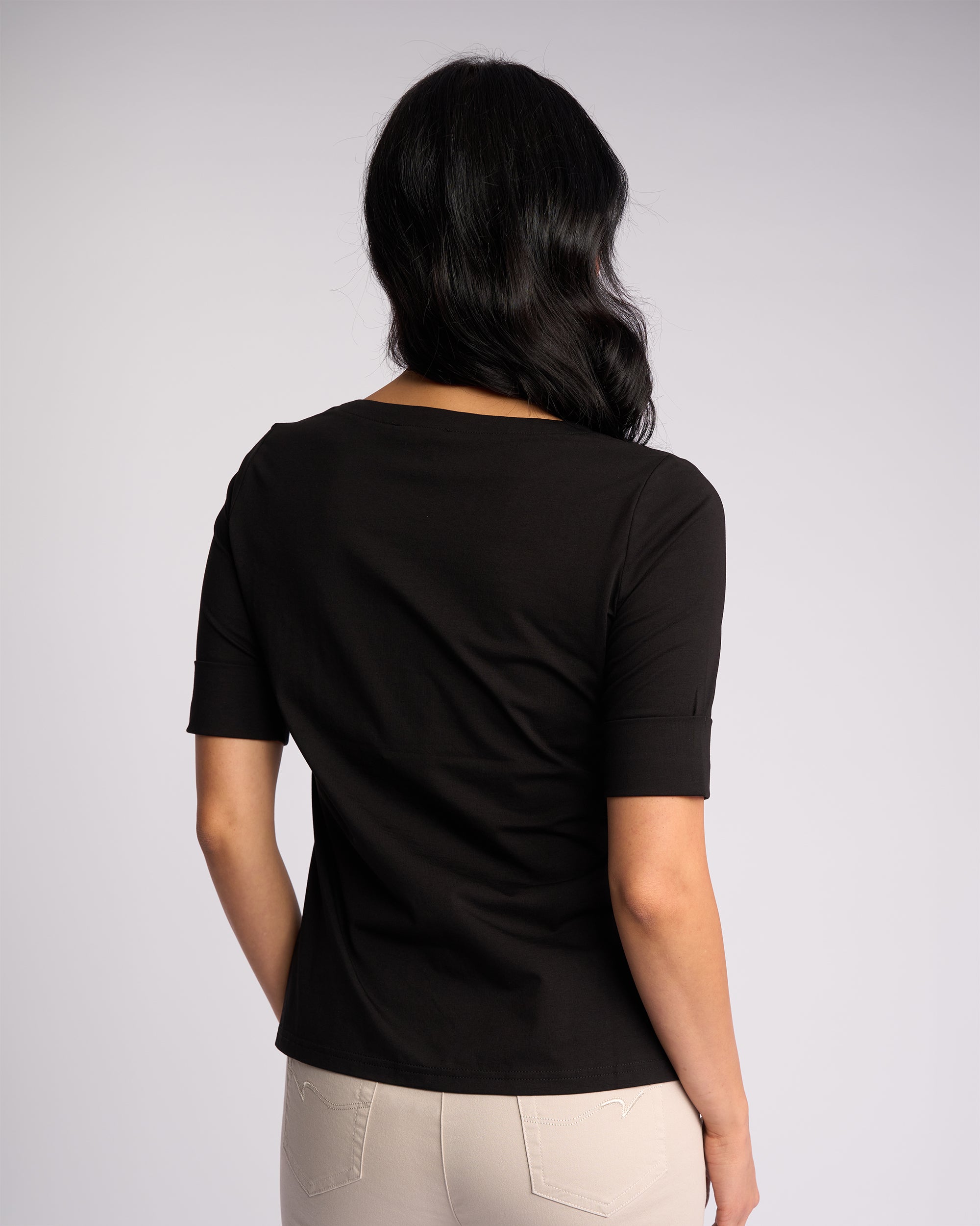 A woman with long dark hair models the Vassalli Reece Top in black, paired with light pants, standing with her back to the camera against a plain backdrop—a stylish and comfortable choice for everyday wear.