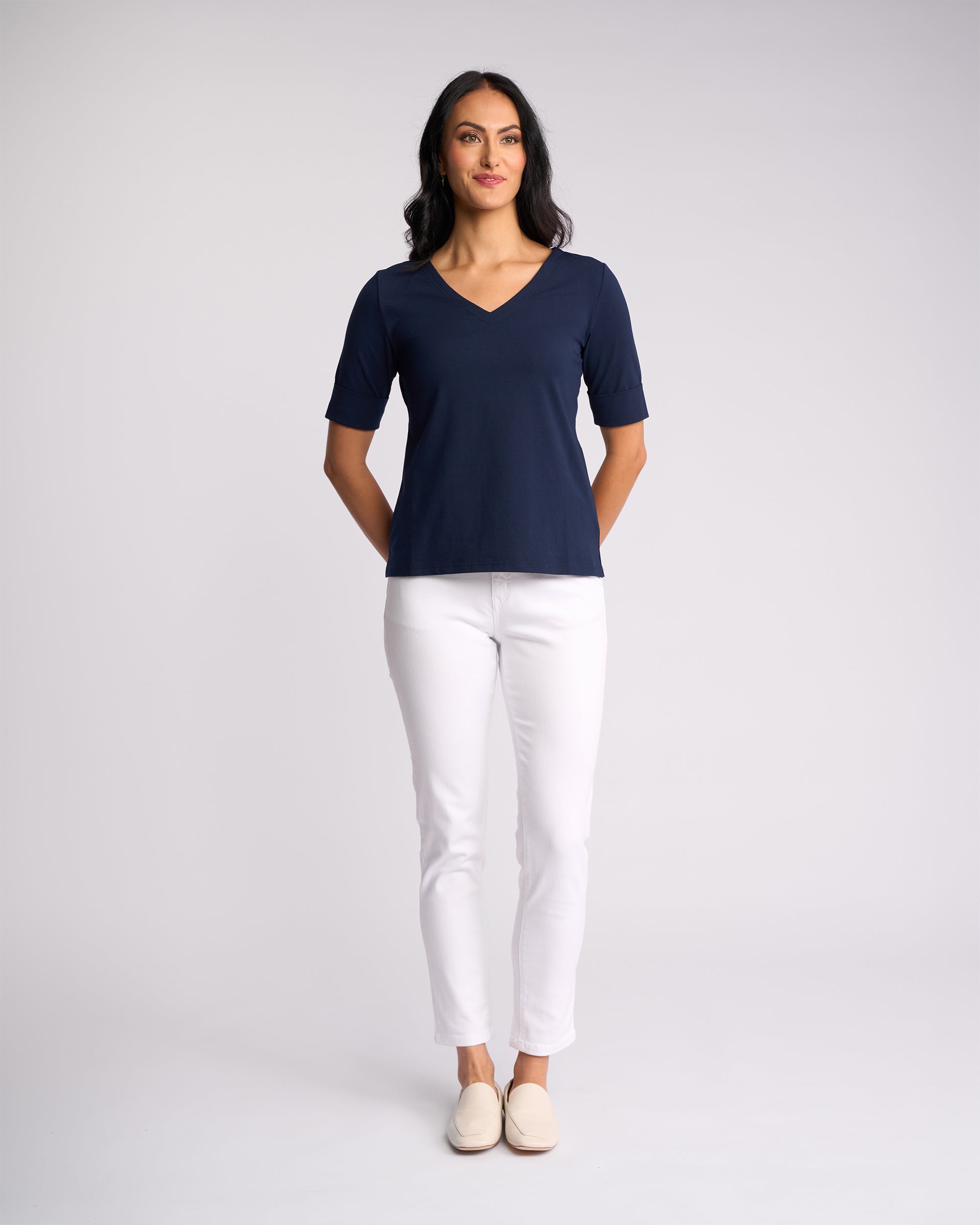 A woman stands against a plain background, smiling slightly, wearing the Vassalli Reece Top - Navy with white pants and beige slip-on shoes. Her hands are behind her back.