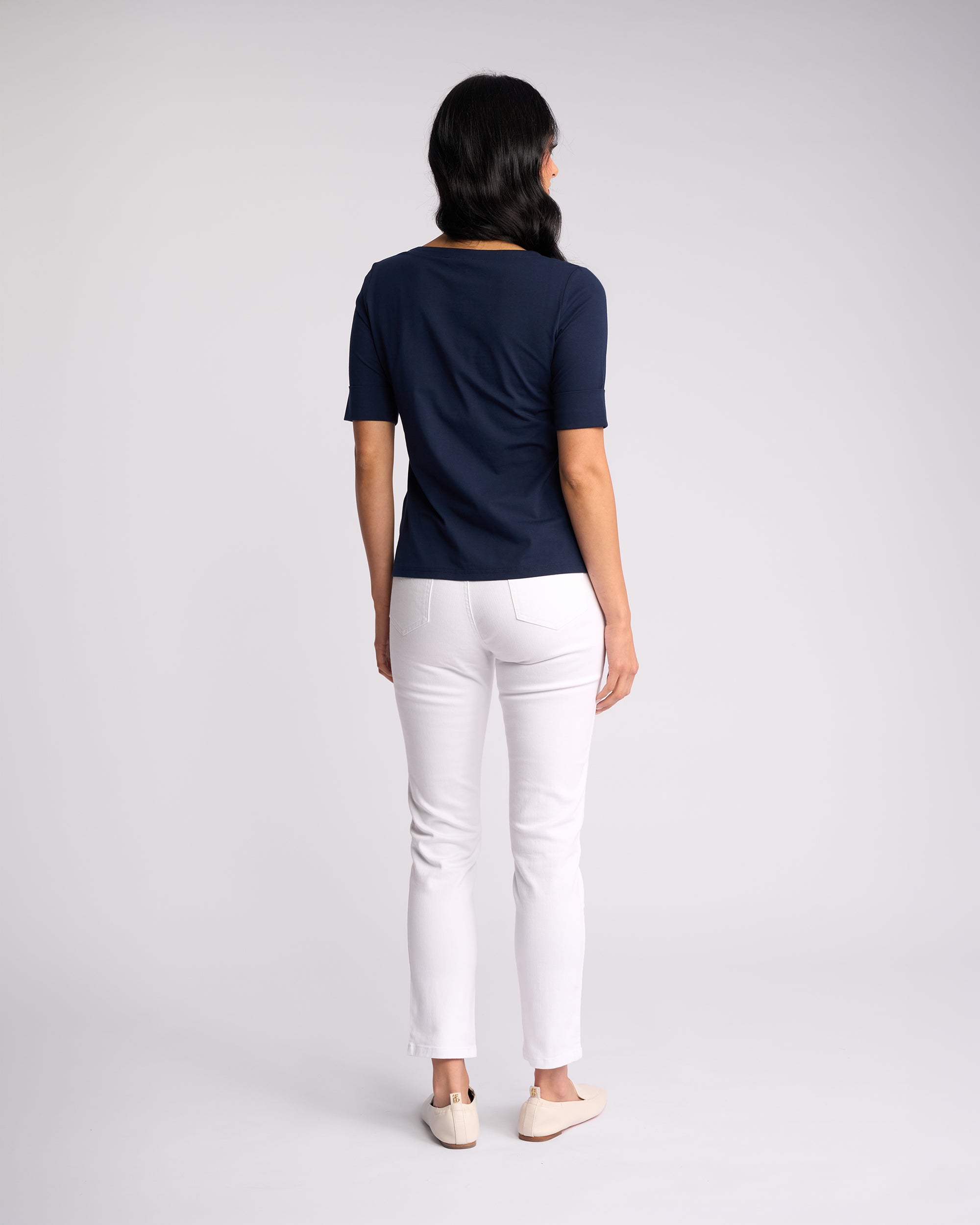 A woman with long dark hair stands facing away, wearing the Vassalli Reece Top - Navy with white pants and beige shoes, set against a plain light gray background.