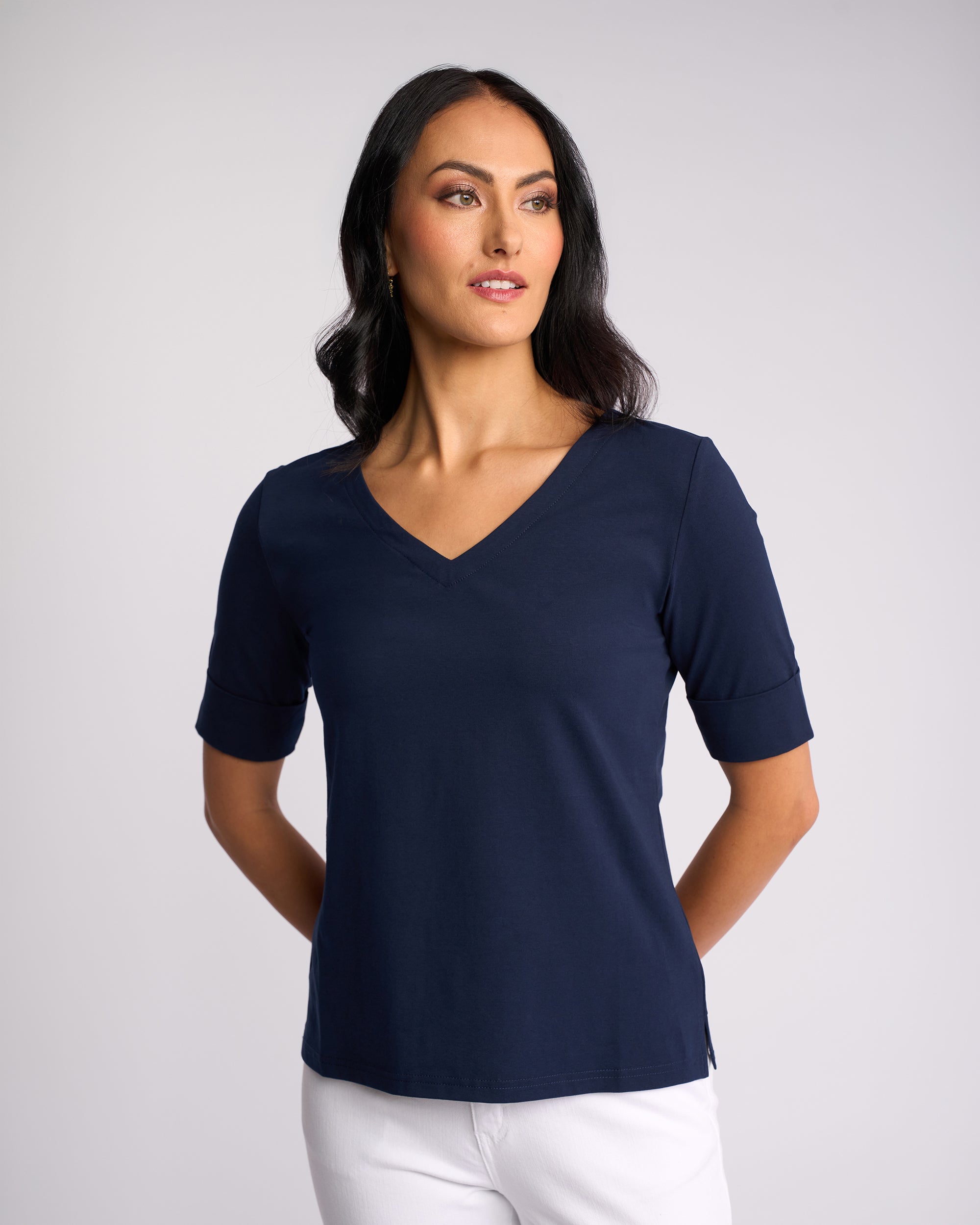 A woman with long dark hair wears the Vassalli Reece Top - Navy, paired with white pants, standing with hands behind her back and looking to the side against a plain light background—ideal for stylish everyday wear.