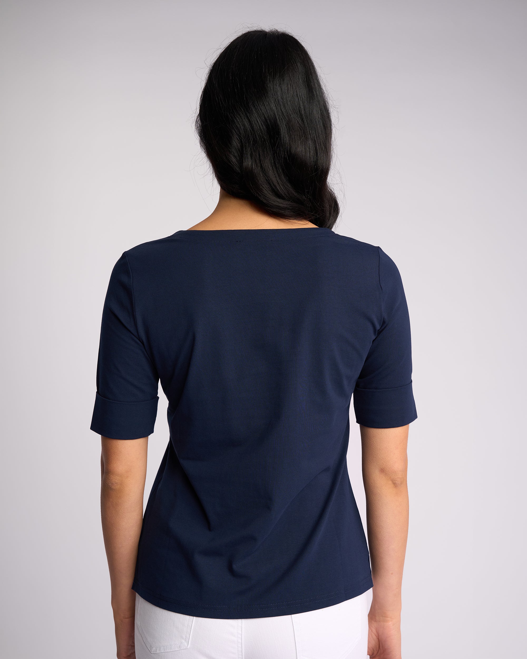 A woman with long dark hair faces away, wearing the Vassalli Reece Top - Navy and white pants, made from soft stretch fabric, set against a plain light gray background.