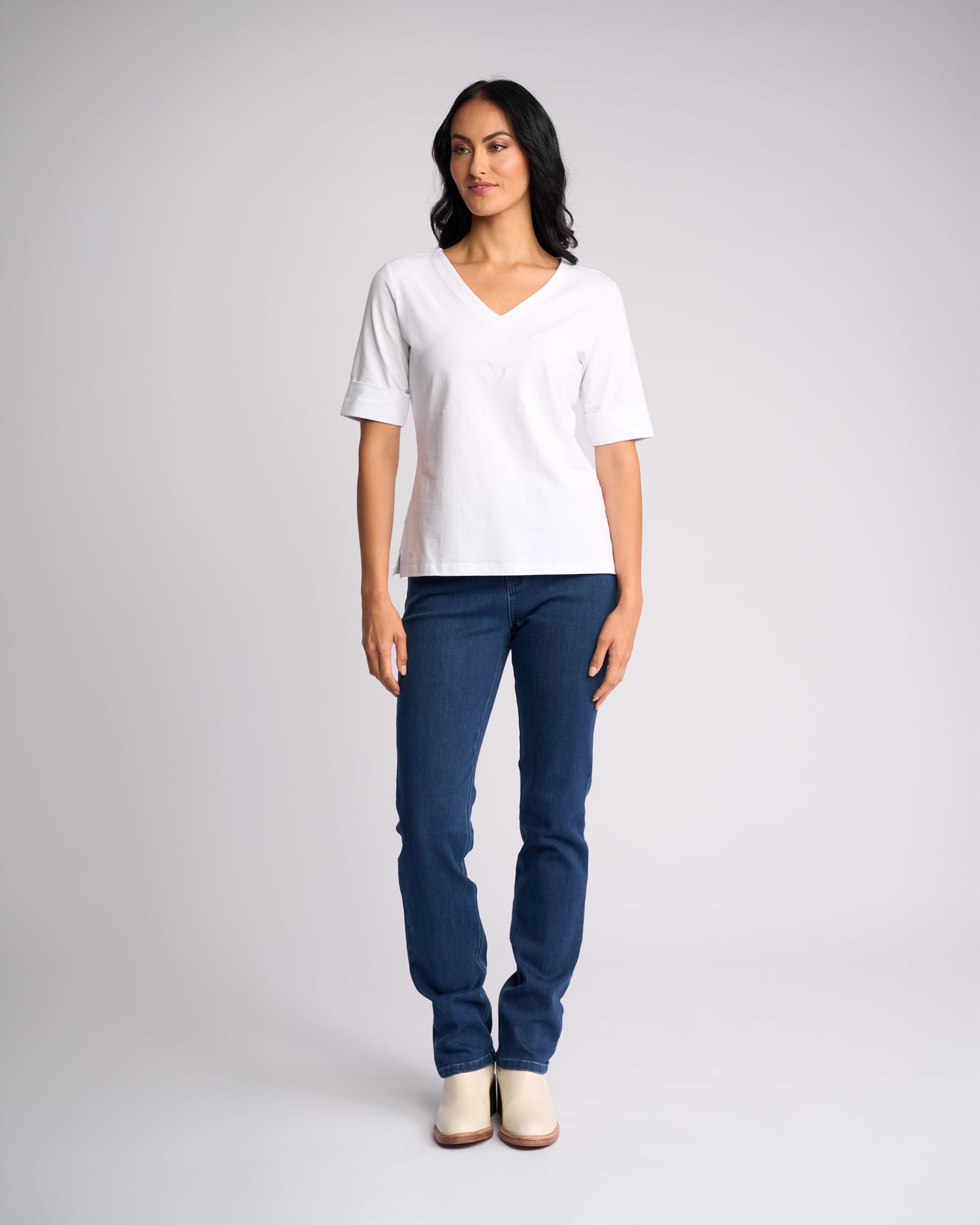 A woman poses against a plain light background wearing the Vassalli Reece Top - White, paired with blue jeans and light-colored shoes. She looks slightly to the side with her arms relaxed at her sides.