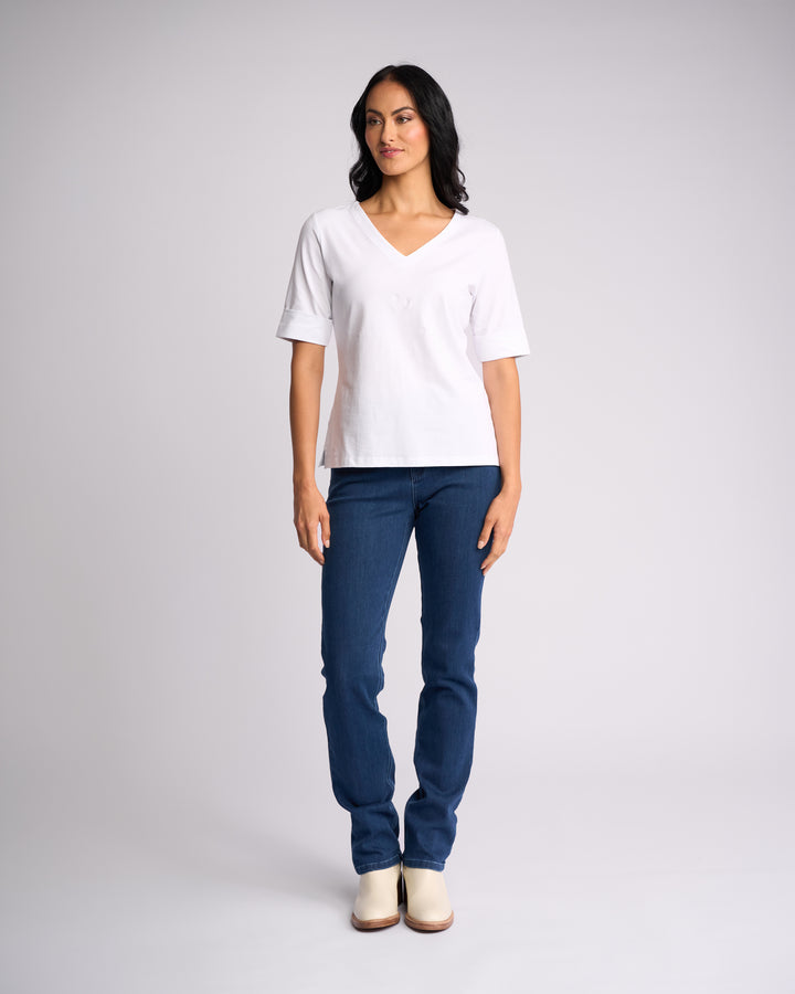 A woman poses against a plain light background wearing the Vassalli Reece Top - White, paired with blue jeans and light-colored shoes. She looks slightly to the side with her arms relaxed at her sides.