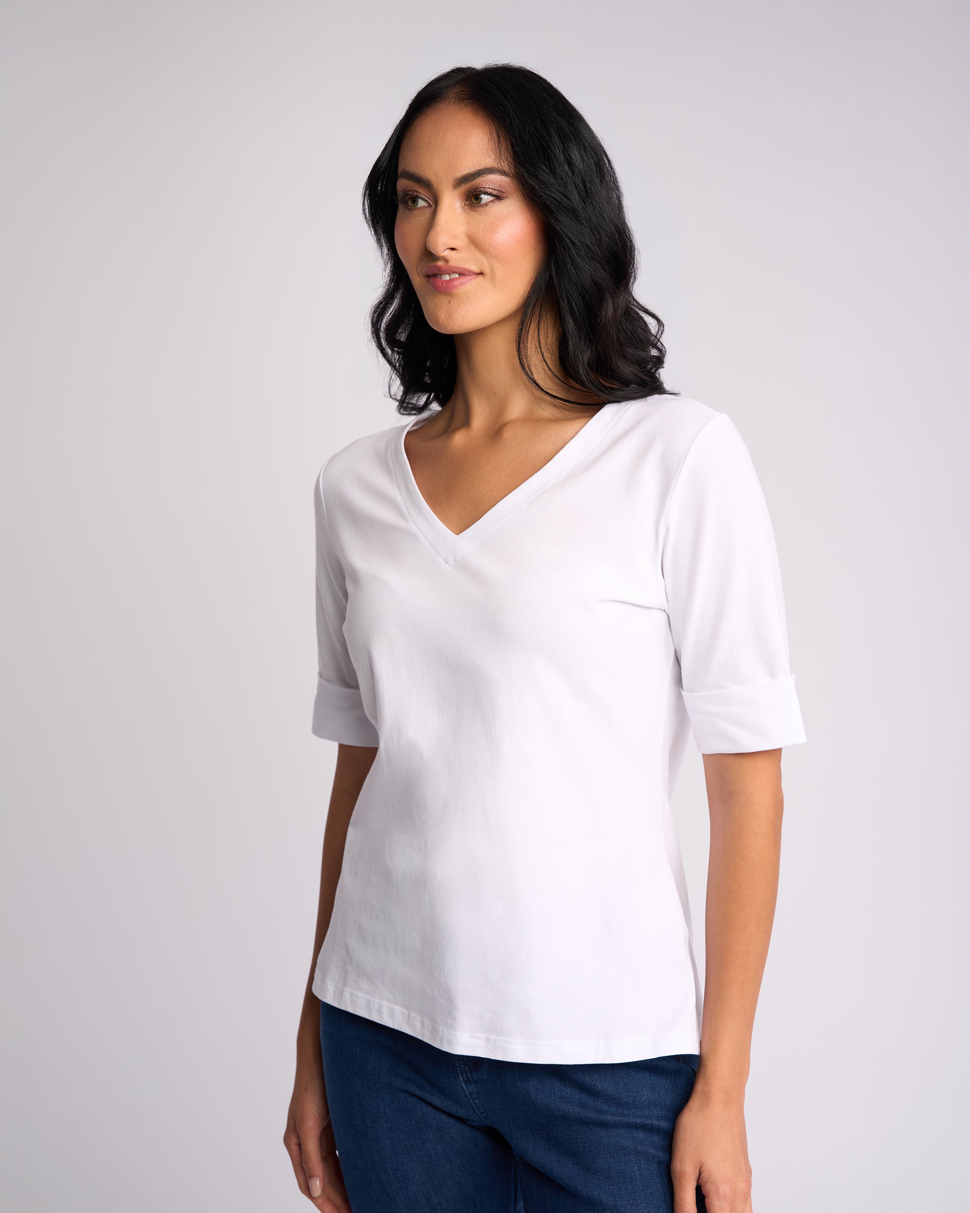 A woman with long dark hair wears the Vassalli Reece Top - White and blue jeans, standing against a plain light background, looking slightly to the side and smiling softly.