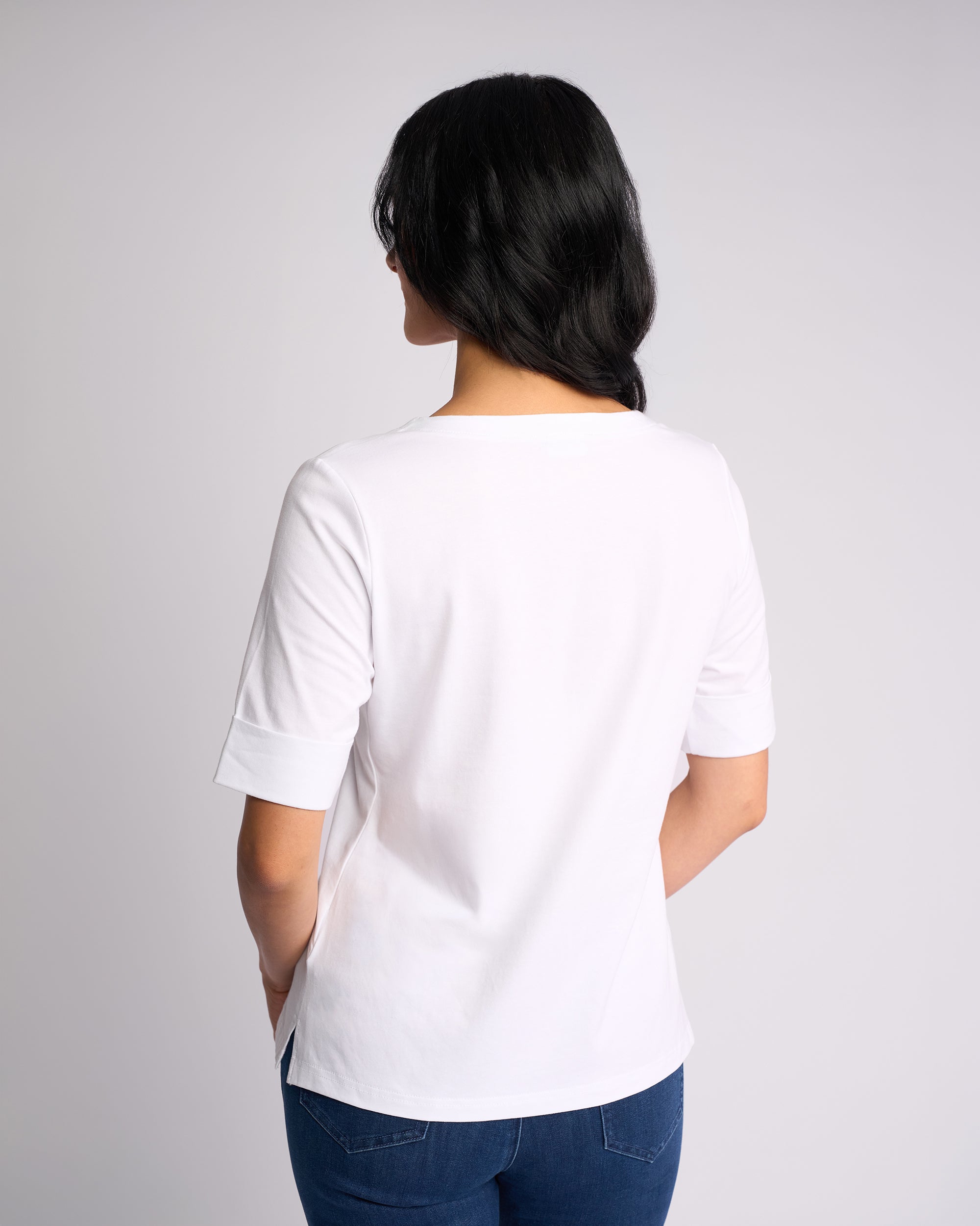 A person with long dark hair, facing away, wears a Vassalli Reece Top - White with stretch fabric and blue jeans against a plain light background.
