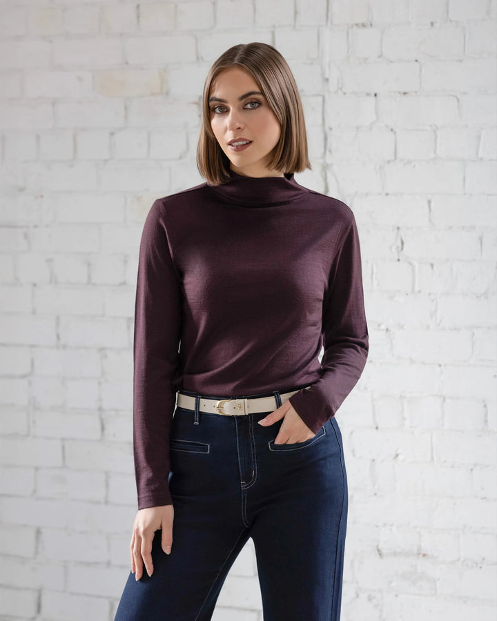 A woman with straight brown hair wears the Vassalli Riley Merino Polo in Berry and dark jeans, standing against a white brick wall. She looks at the camera with a slight smile, one hand in her pocket.