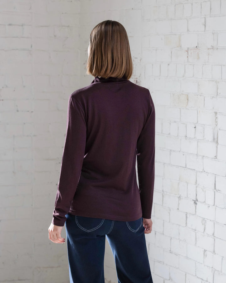 A person with straight, shoulder-length brown hair faces a white brick wall, wearing the Vassalli Riley Merino Polo in Berry and dark blue jeans. Their back is to the camera.