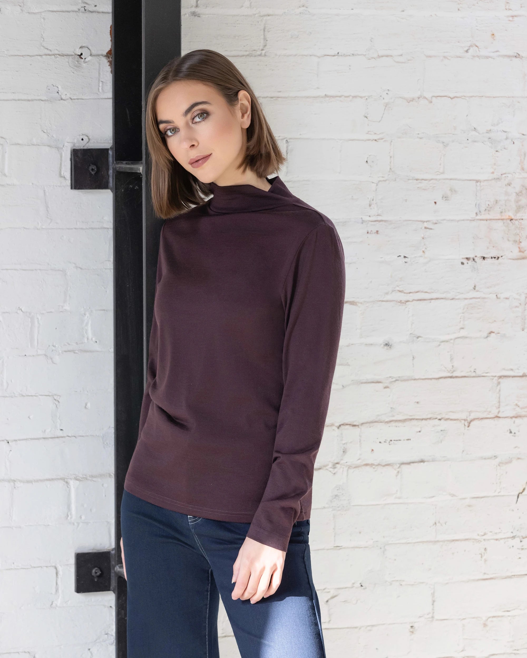 A woman with straight brown hair, wearing the Vassalli Riley Merino Polo in Berry and blue jeans, stands against a white brick wall in natural light, calmly looking at the camera.