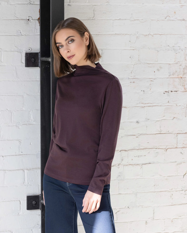 A woman with straight brown hair, wearing the Vassalli Riley Merino Polo in Berry and blue jeans, stands against a white brick wall in natural light, calmly looking at the camera.