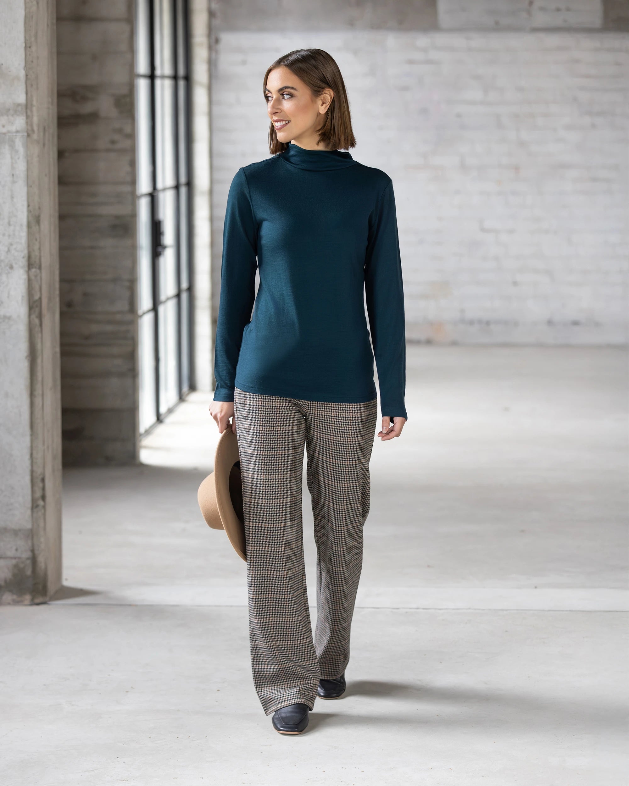 Wearing the Vassalli Riley Merino Polo in Dragonfly, a smiling woman with short brown hair walks indoors, holding a tan hat by her side. Large windows and a concrete wall are in the background.