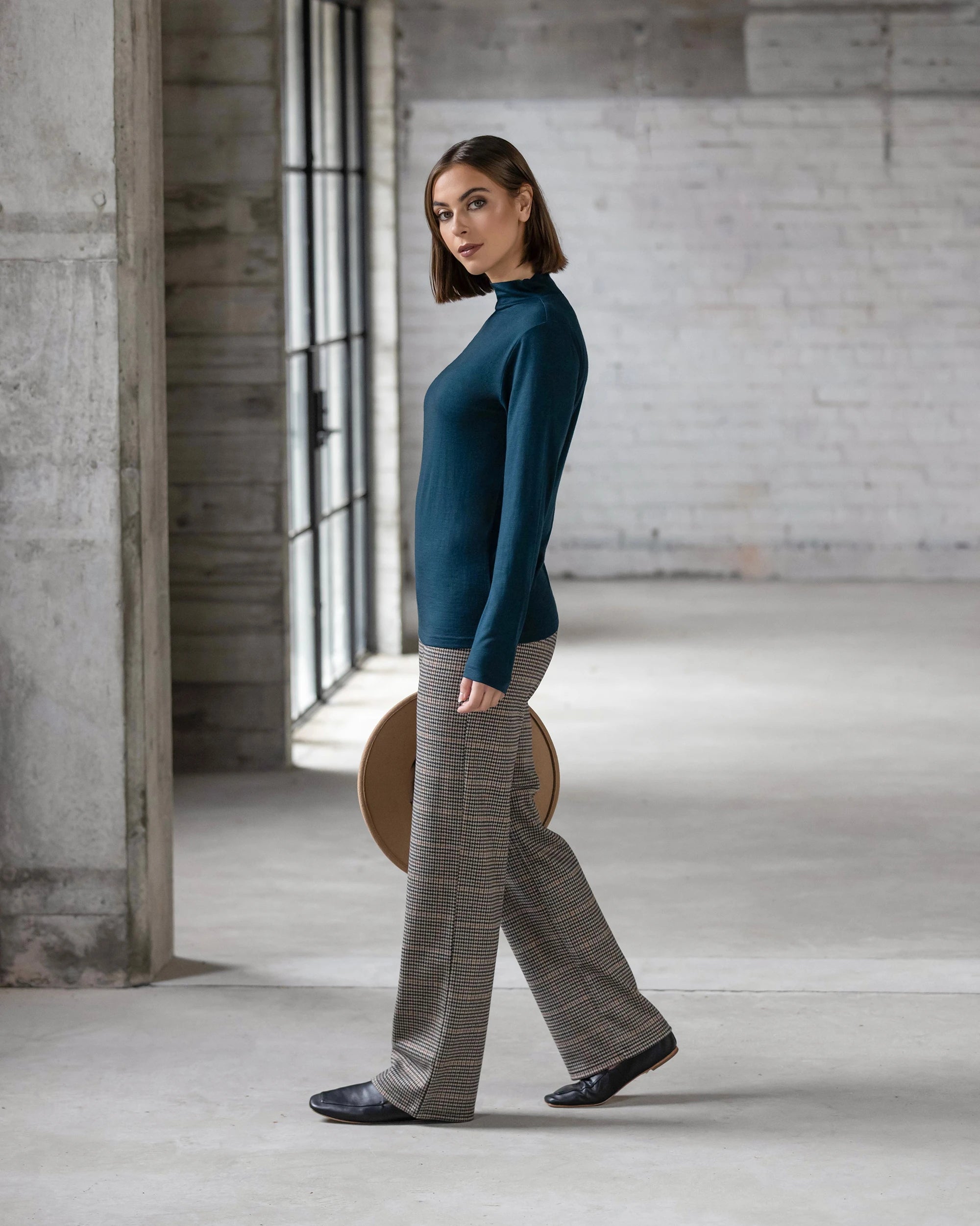 A woman with shoulder-length brown hair, wearing the Vassalli Riley Merino Polo in Dragonfly and plaid trousers, stands sideways in a spacious, light-filled industrial room with concrete floors and brick walls.