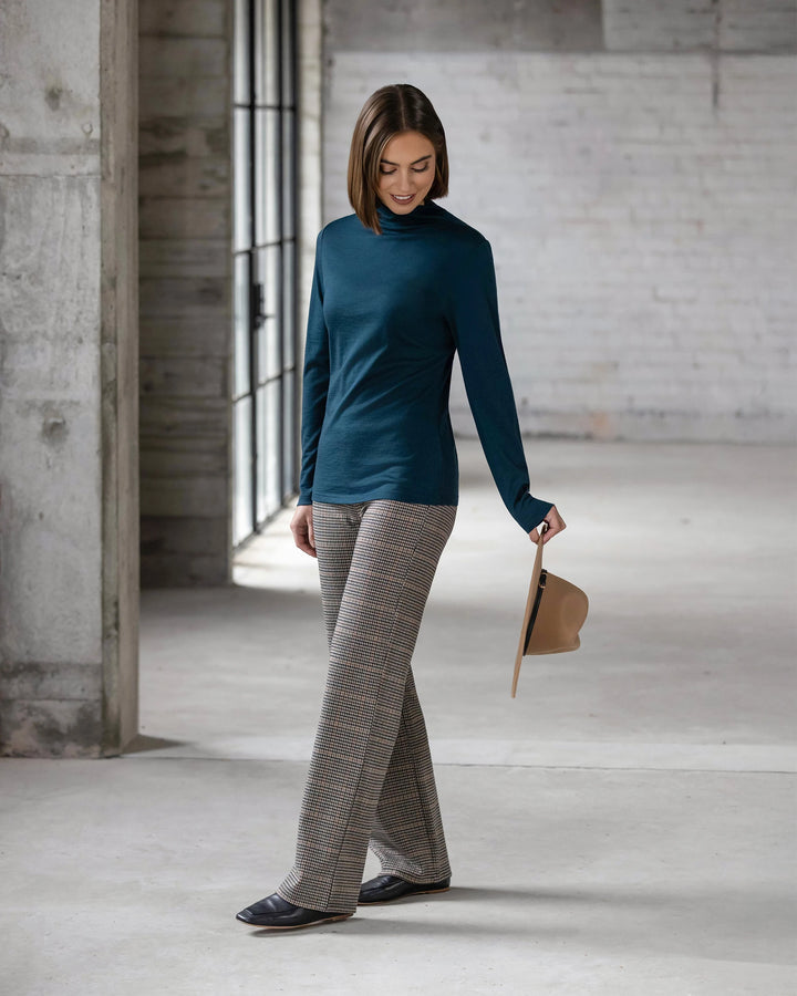 A woman stands indoors on a concrete floor, wearing the Vassalli Riley Merino Polo - Dragonfly with plaid trousers and black shoes. She holds a tan hat in one hand while looking down as light streams through tall windows.
