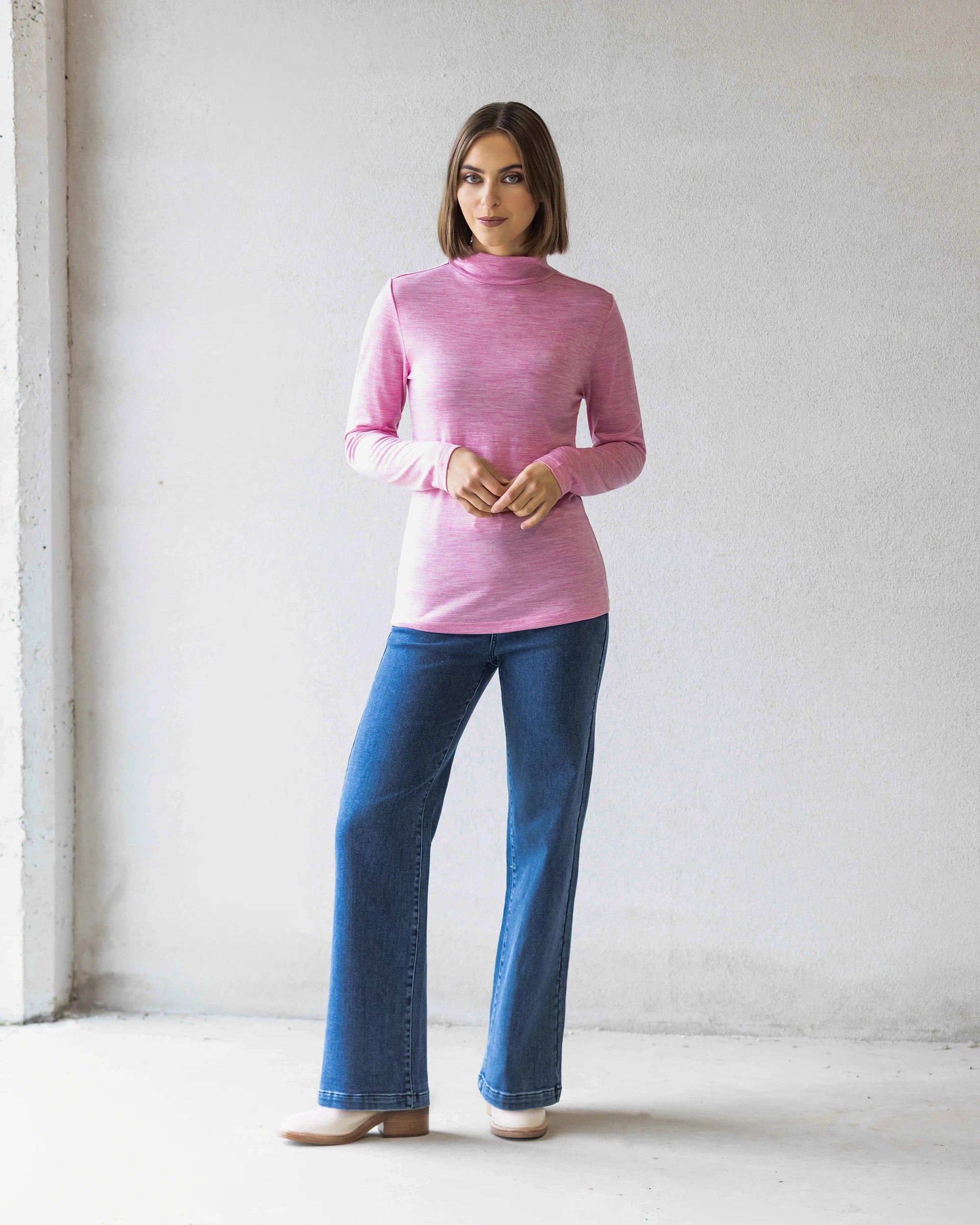 A woman stands indoors against a light wall, wearing the Vassalli Riley Merino Polo in Pink Marle with blue jeans and beige shoes. She faces forward with her hands clasped—a perfect winter layer.
