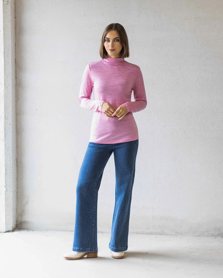 A woman stands indoors against a light wall, wearing the Vassalli Riley Merino Polo in Pink Marle with blue jeans and beige shoes. She faces forward with her hands clasped—a perfect winter layer.