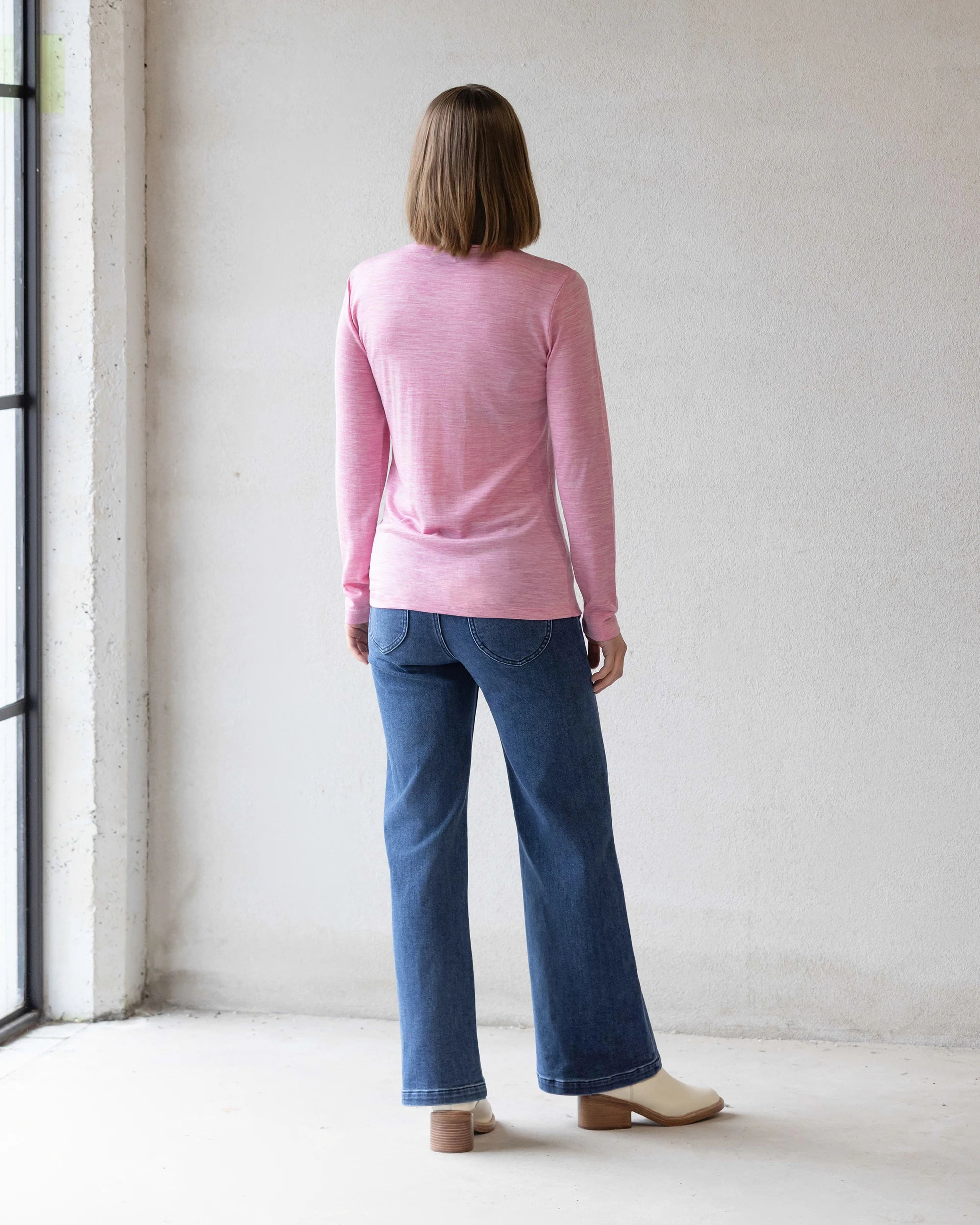 A person with shoulder-length hair, wearing the Vassalli Riley Merino Polo in Pink Marle, blue jeans, and cream shoes stands facing a white wall with their back to the camera.