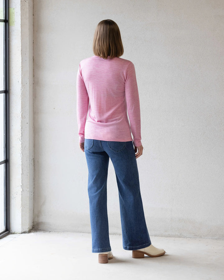 A person with shoulder-length hair, wearing the Vassalli Riley Merino Polo in Pink Marle, blue jeans, and cream shoes stands facing a white wall with their back to the camera.