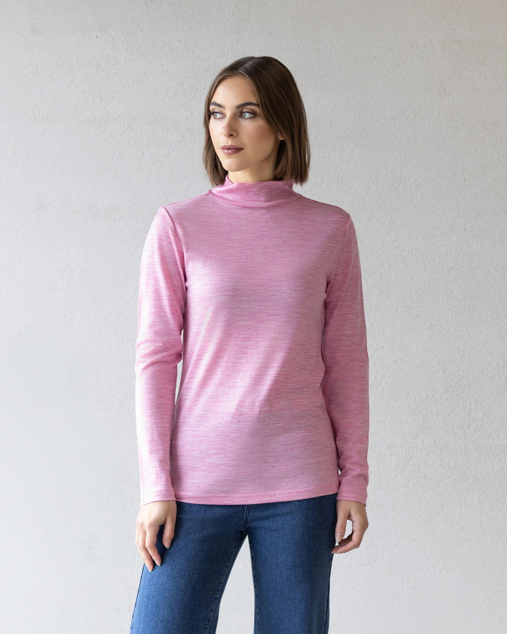 A woman with straight brown hair wears the Vassalli Riley Merino Polo in Pink Marle with blue jeans, standing against a light gray textured wall and looking slightly to her left.