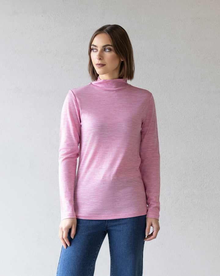 A woman with straight brown hair wears the Vassalli Riley Merino Polo in Pink Marle with blue jeans, standing against a light gray textured wall and looking slightly to her left.