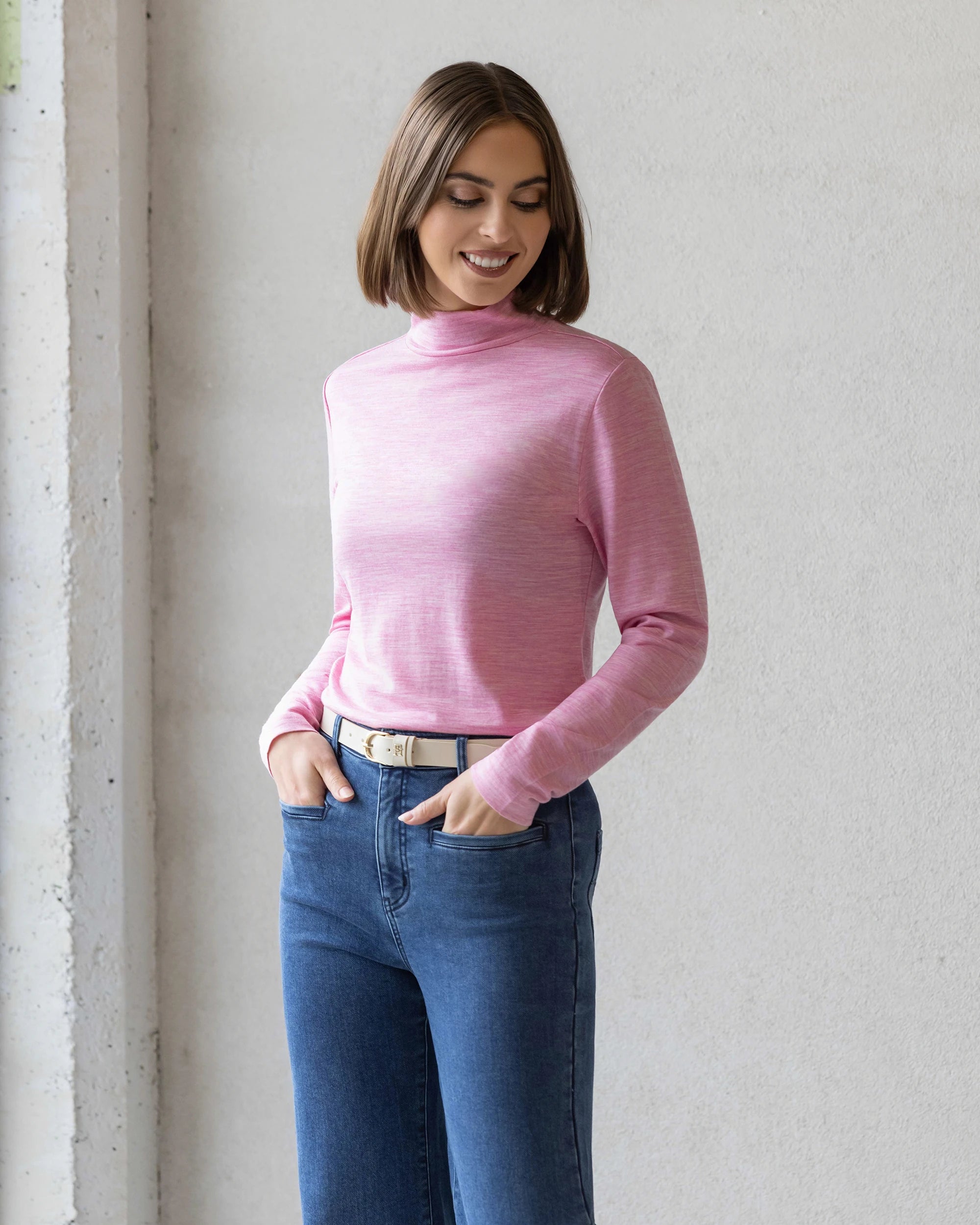A woman with a bob haircut smiles with her hands in her pockets, wearing the Vassalli Riley Merino Polo in Pink Marle and blue jeans, standing against a light-colored wall.