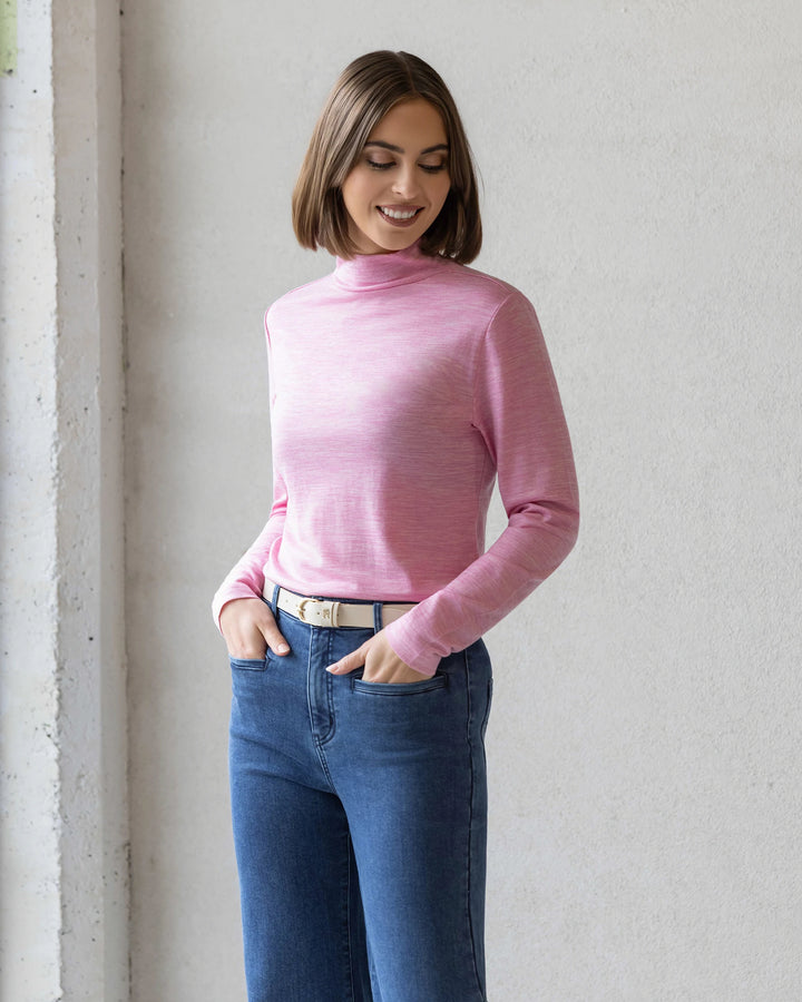 A woman with a bob haircut smiles with her hands in her pockets, wearing the Vassalli Riley Merino Polo in Pink Marle and blue jeans, standing against a light-colored wall.