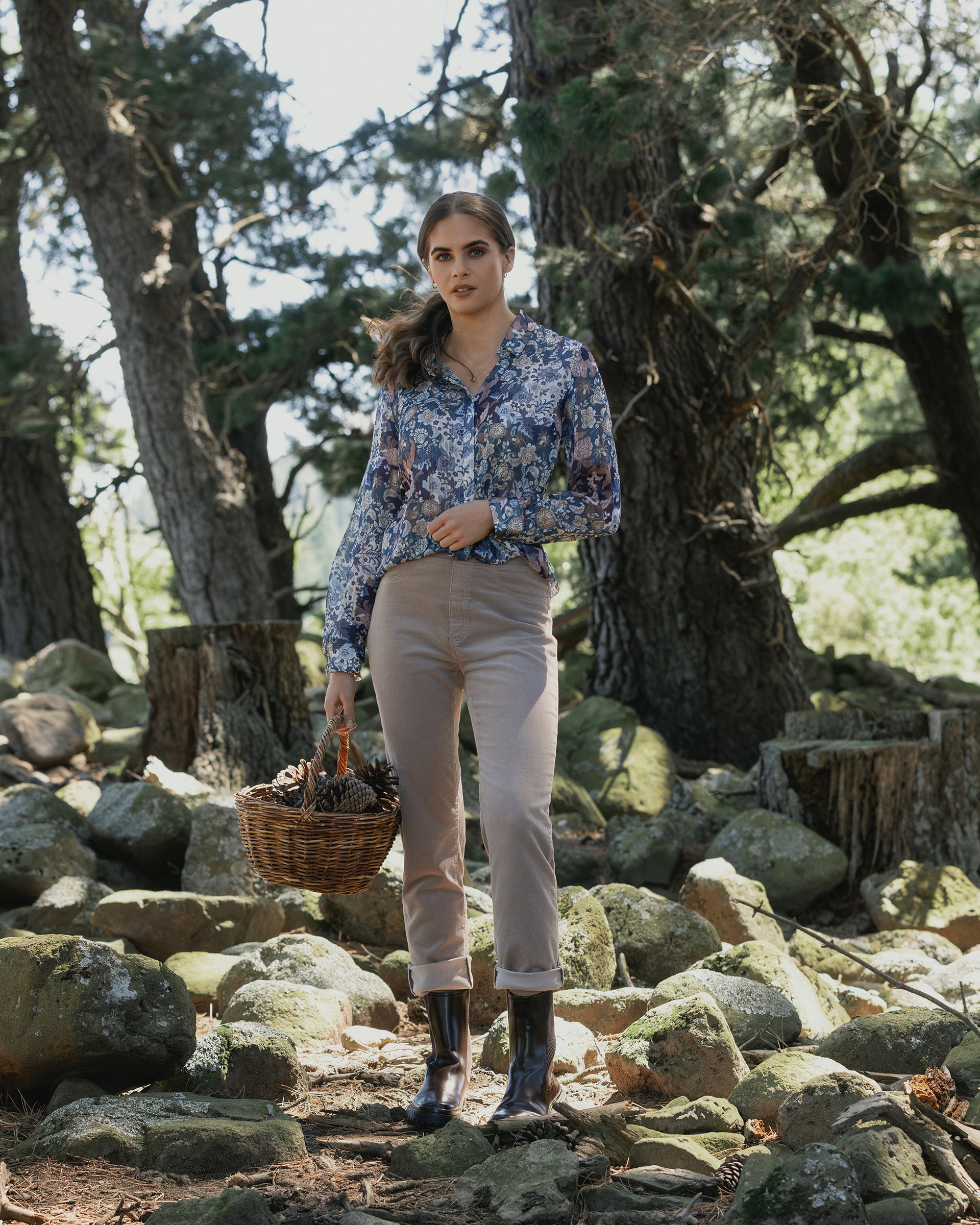 A woman in a floral shirt, Vassalli Bay Cord Pull On - Twig pants, and black boots stands on rocky ground in a forest, holding a basket of pinecones as sunlight filters through the trees.