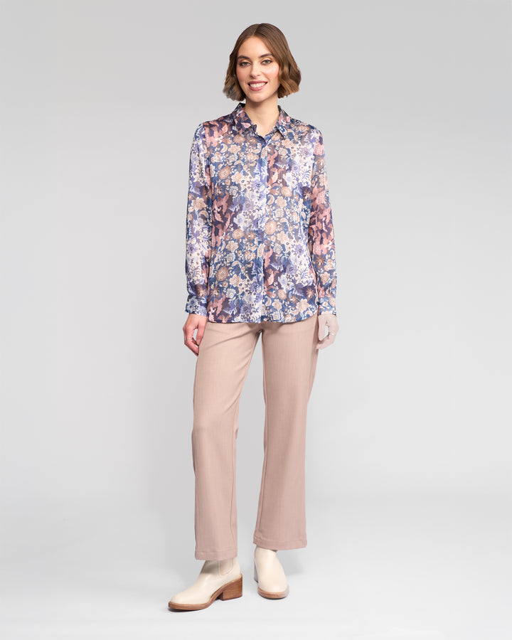 A woman smiles in Vassalli's Ballot Shirt - Rhea, a long-sleeve floral blouse in blue and pink, paired with beige pants and cream ankle boots against a plain light gray background.