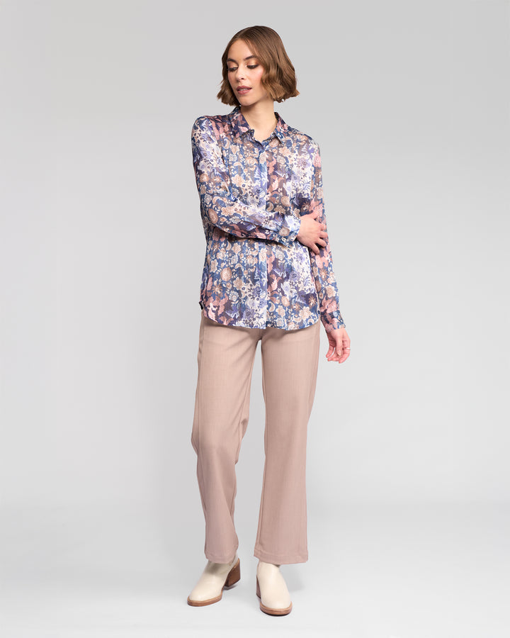 A woman with short, wavy hair looks down, gently touching her arm as she models the Vassalli Ballot Shirt - Rhea against a plain background, paired with beige pants and light ankle boots.