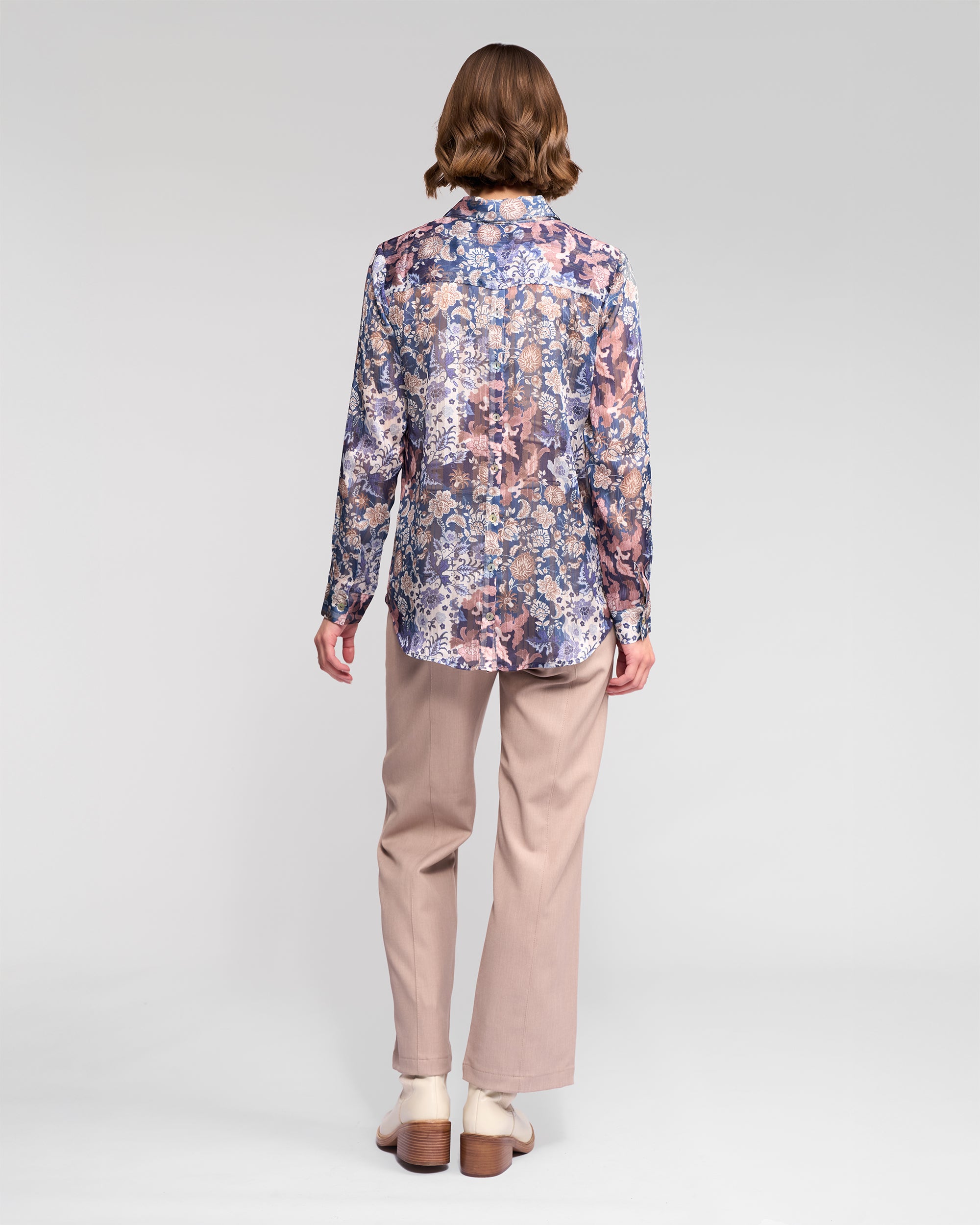 A person stands facing away, wearing the Vassalli Ballot Shirt - Rhea, paired with light beige pants and white shoes against a plain light gray background.