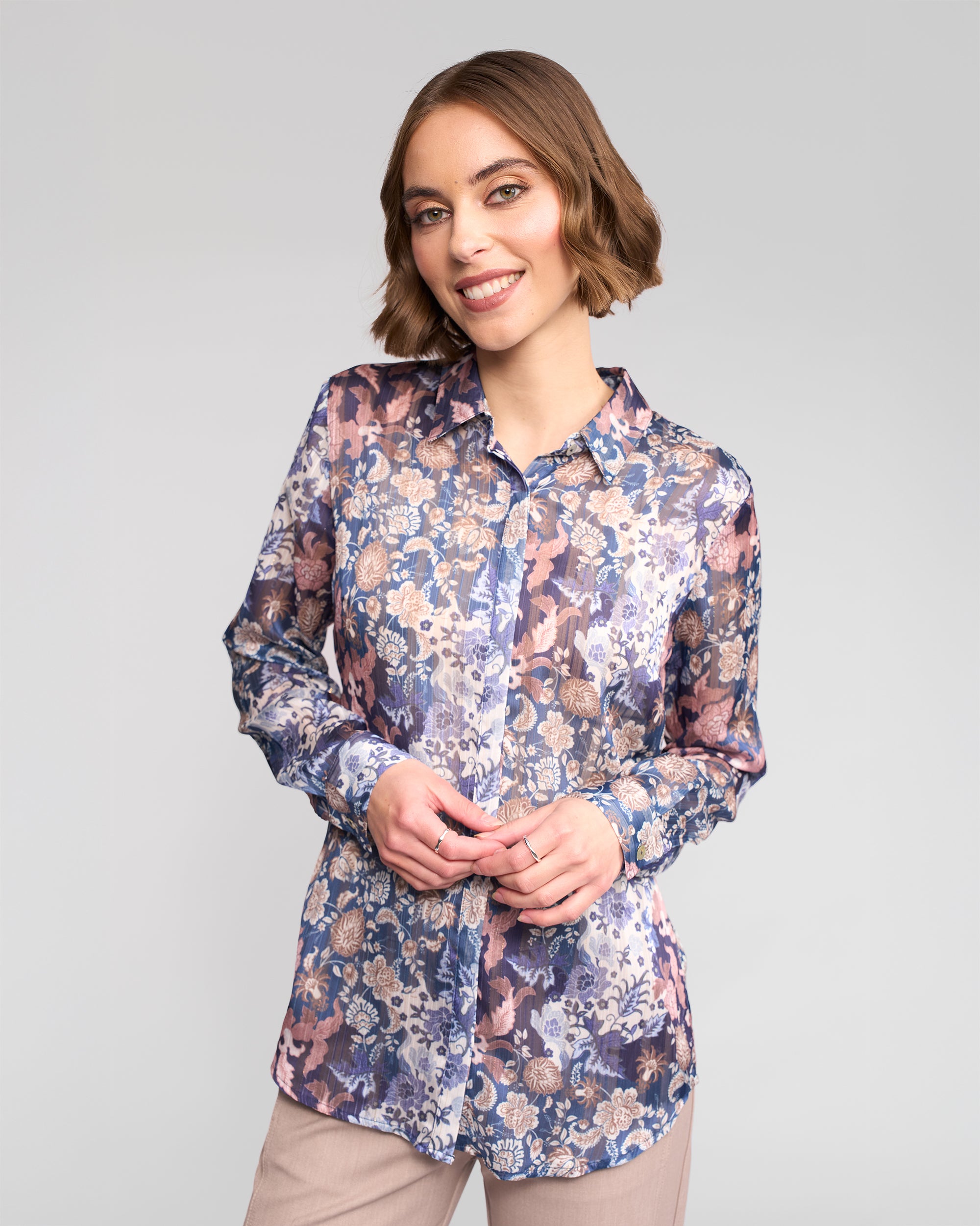 A woman with short brown hair smiles against a light gray background, wearing the Vassalli Ballot Shirt - Rhea, a long-sleeve women’s top with a purple, blue, and beige floral pattern, paired with light-colored pants.