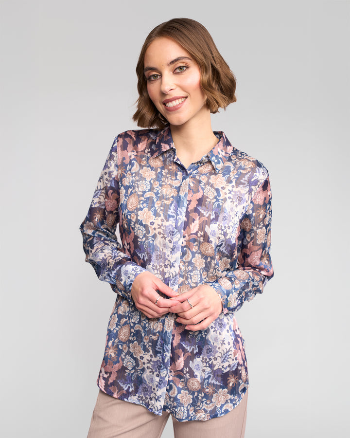 A woman with short brown hair smiles against a light gray background, wearing the Vassalli Ballot Shirt - Rhea, a long-sleeve women’s top with a purple, blue, and beige floral pattern, paired with light-colored pants.
