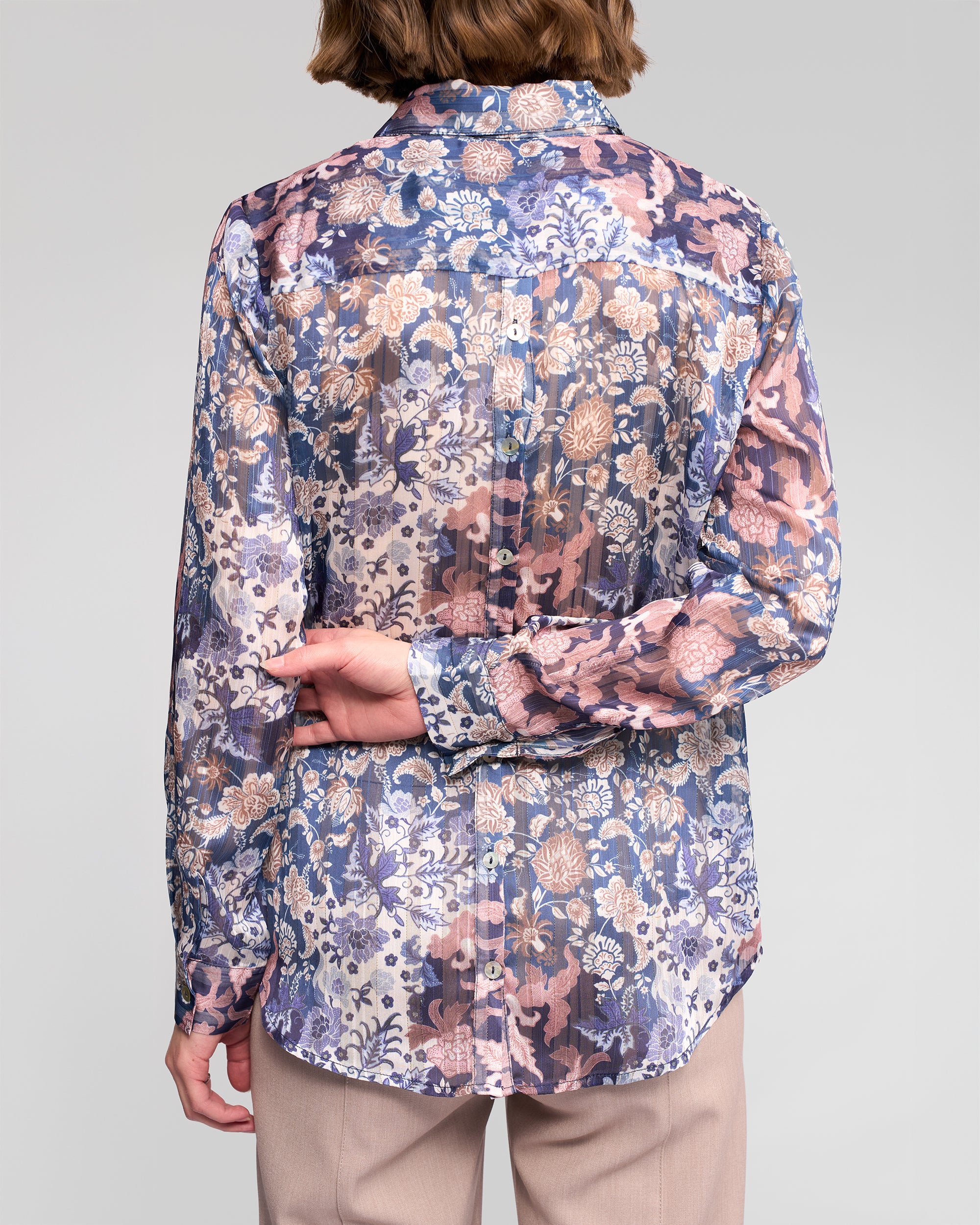 A person is shown from the back wearing Vassalli's Ballot Shirt - Rhea, a women's long-sleeve floral chiffon blouse in blue, pink, and beige tones with tan pants. The sheer shirt complements the pose with one hand behind their back holding the other wrist.