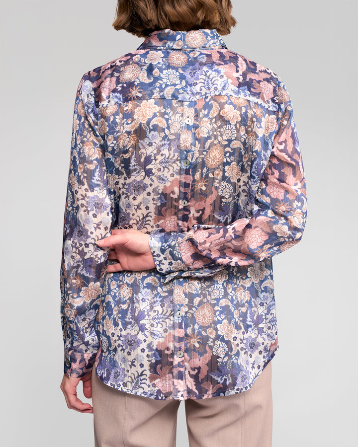 A person is shown from the back wearing Vassalli's Ballot Shirt - Rhea, a women's long-sleeve floral chiffon blouse in blue, pink, and beige tones with tan pants. The sheer shirt complements the pose with one hand behind their back holding the other wrist.
