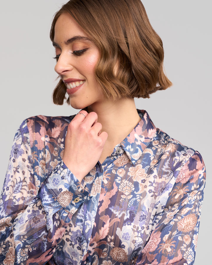 A woman with short, wavy brown hair smiles while looking down, lightly touching her chin. She wears the Vassalli Ballot Shirt - Rhea floral blouse in blue, pink, and beige against a light gray background.