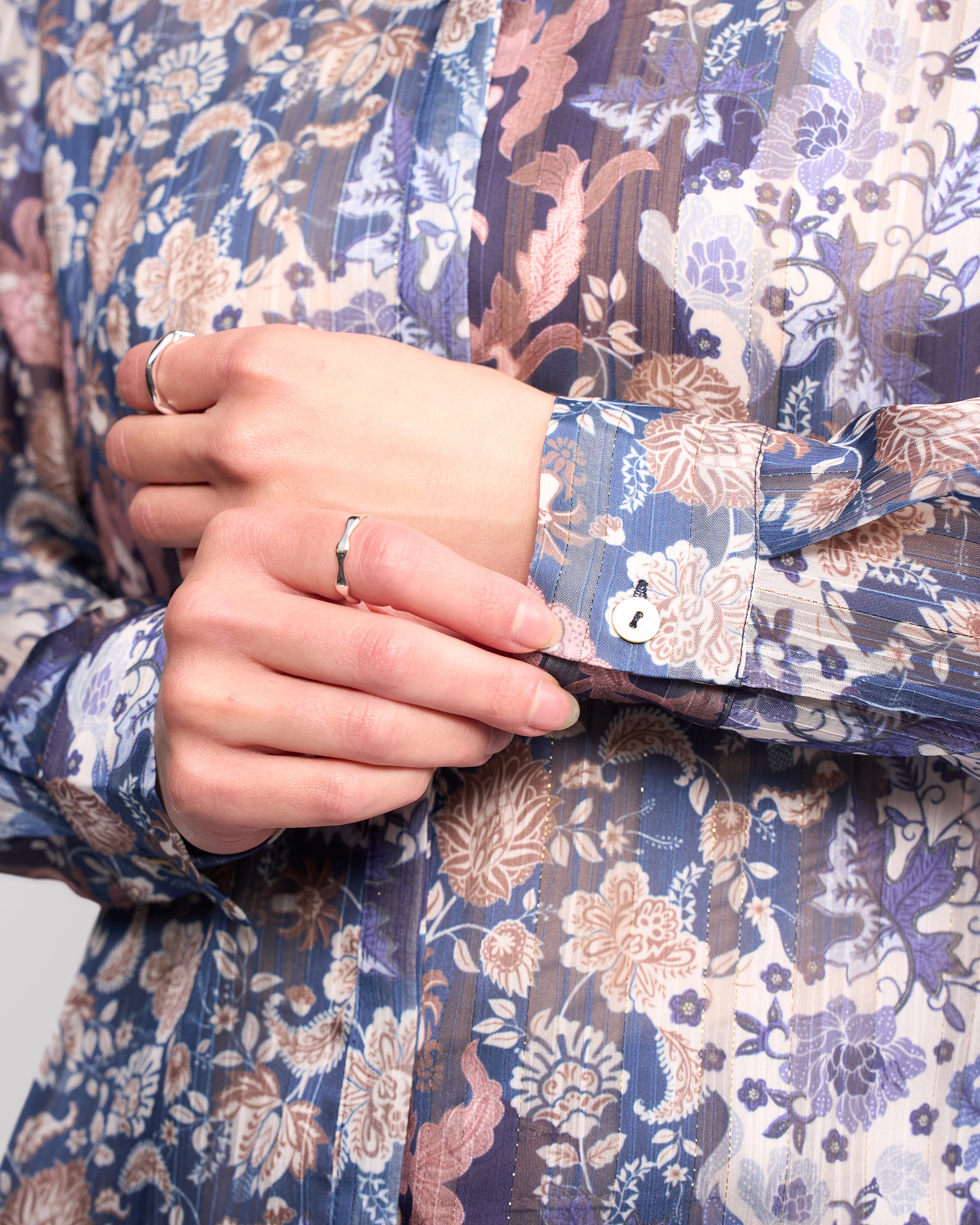 A person wears the Vassalli Ballot Shirt - Rhea, a sheer floral women's blouse in blue, brown, and cream tones, with long sleeves. Their hands are gently clasped together and adorned with silver rings.