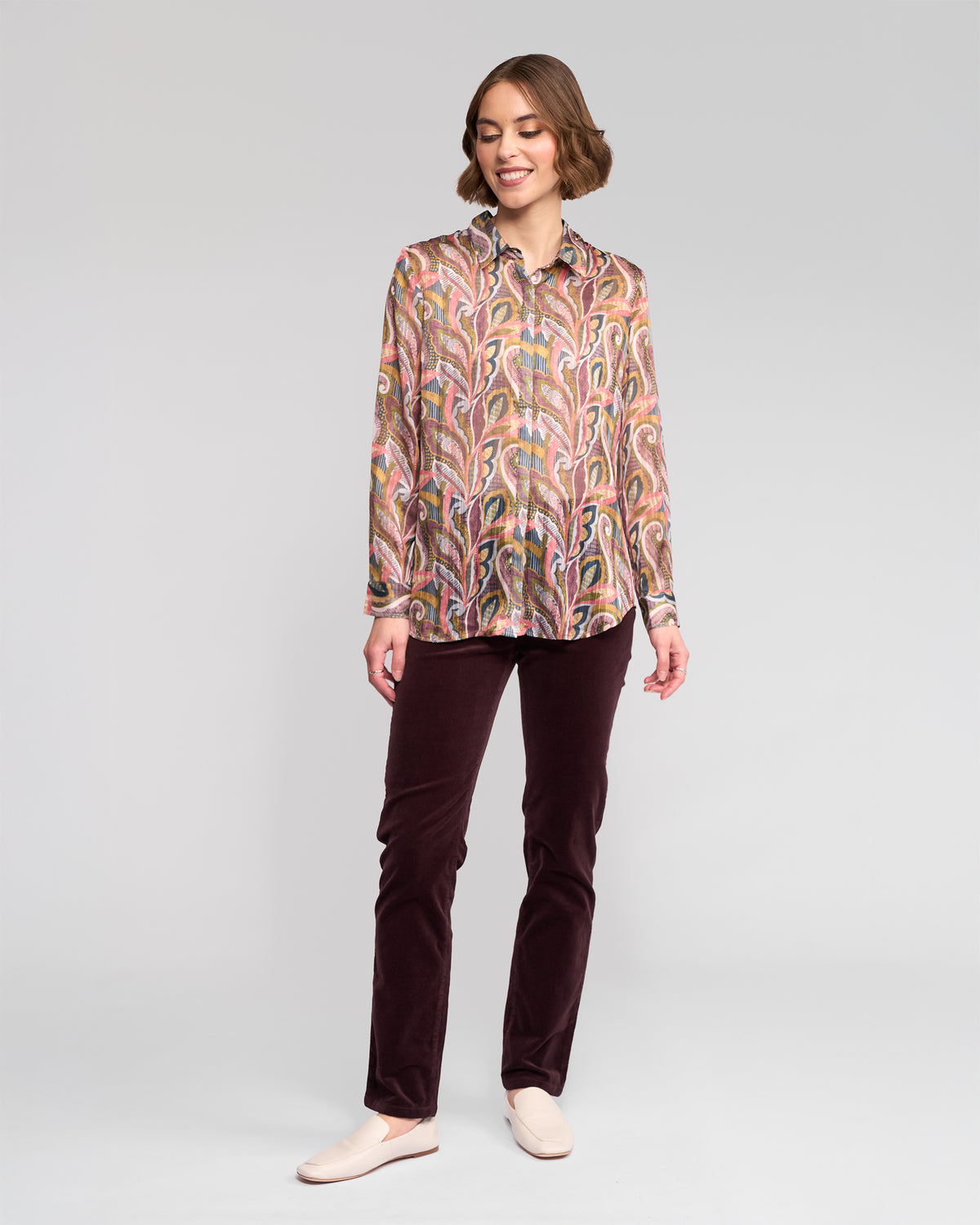 A woman stands smiling, wearing the Vassalli Ballot Shirt - Sable with abstract paisley print, dark purple trousers, and light slip-on shoes against a plain light gray background.