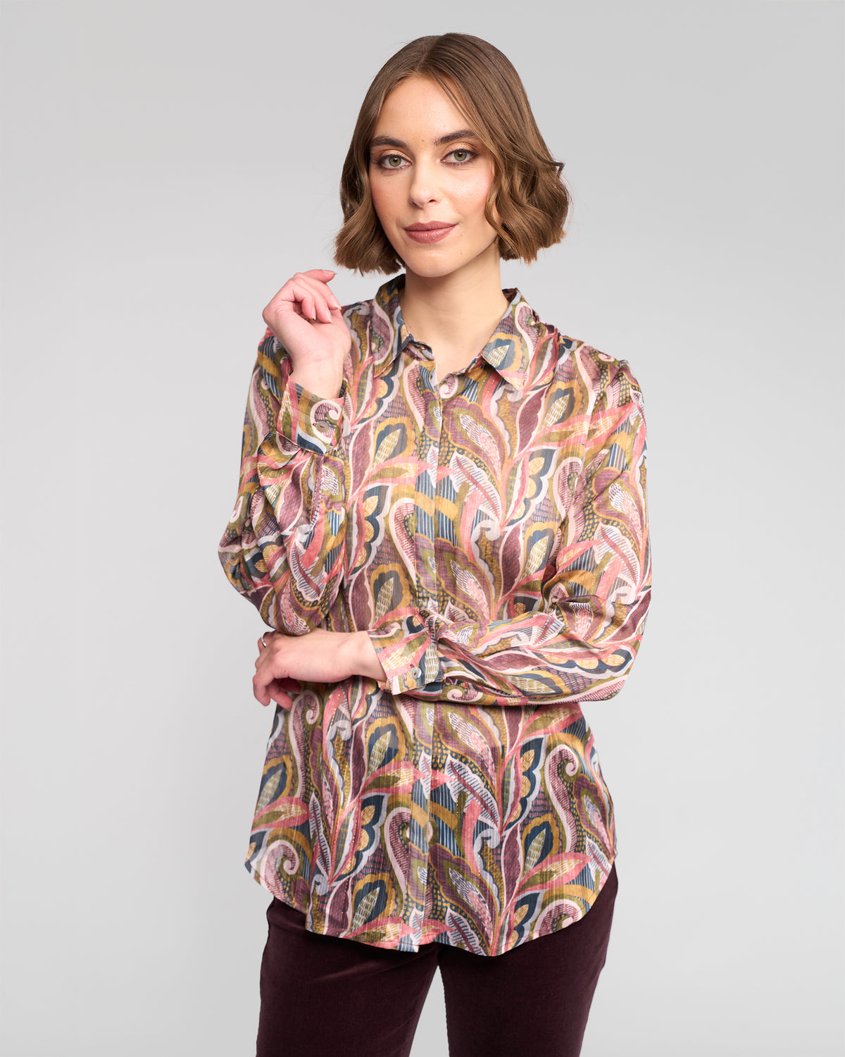 A woman with short brown hair models the Vassalli Ballot Shirt - Sable, standing against a plain light gray background. She wears dark pants and faces the camera with a slight smile, one hand raised near her face.