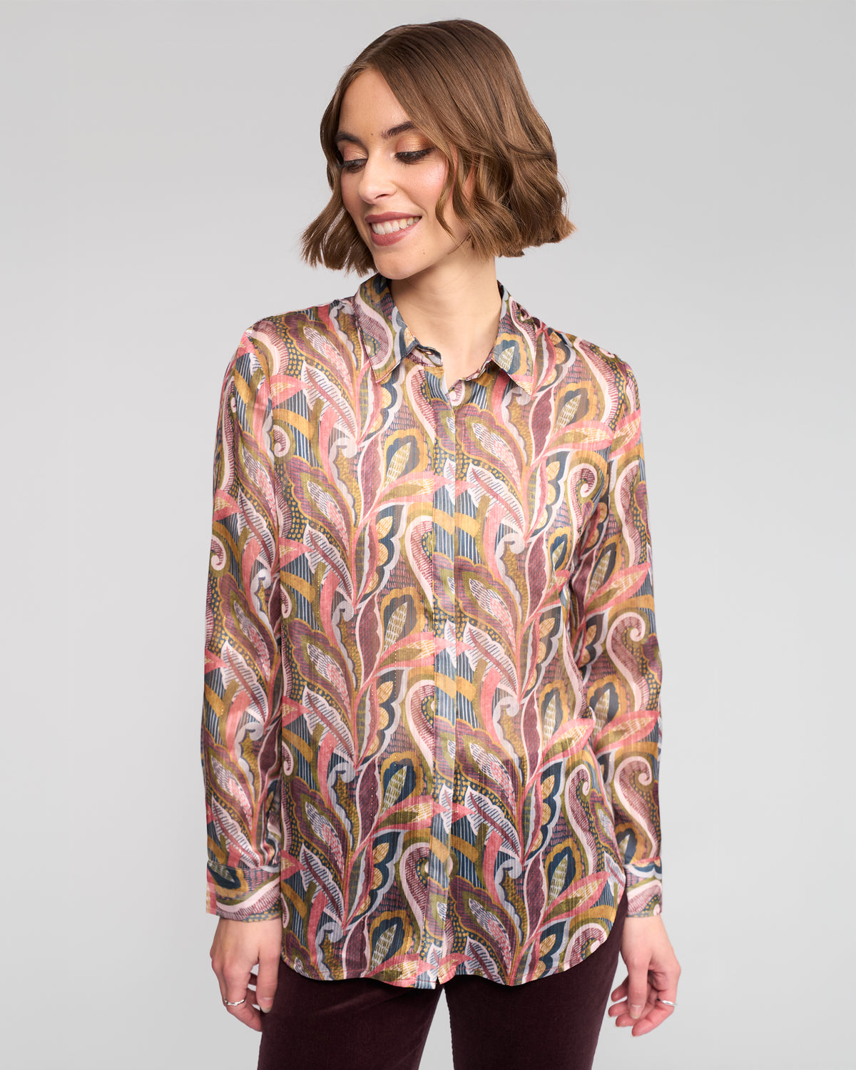 A woman with short brown hair smiles while wearing the Vassalli Ballot Shirt - Sable, a long-sleeve paisley print women’s top with leaf-like designs, paired with dark pants against a plain light gray background.