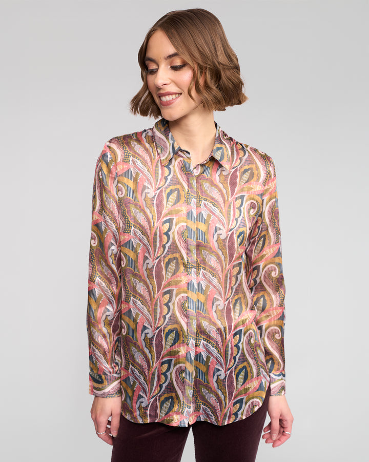 A woman with short brown hair smiles while wearing the Vassalli Ballot Shirt - Sable, a long-sleeve paisley print women’s top with leaf-like designs, paired with dark pants against a plain light gray background.
