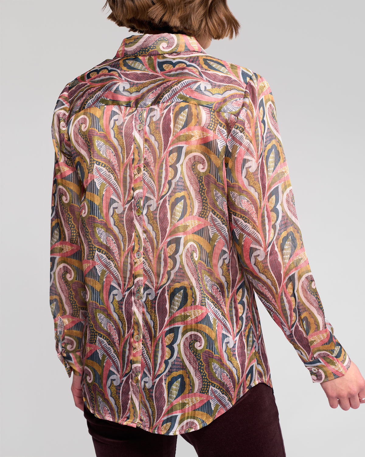 A person is seen from the back wearing the Vassalli Ballot Shirt - Sable, a long-sleeve women's top featuring a colorful paisley pattern with swirling red, yellow, green, and white designs.