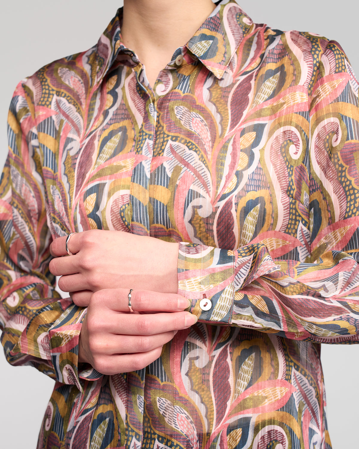 A person wearing the Vassalli Ballot Shirt - Sable, featuring long sleeves with pink, yellow, and green patterns, adjusts their cuff. Their torso and hands are visible, accessorized with several thin silver rings.