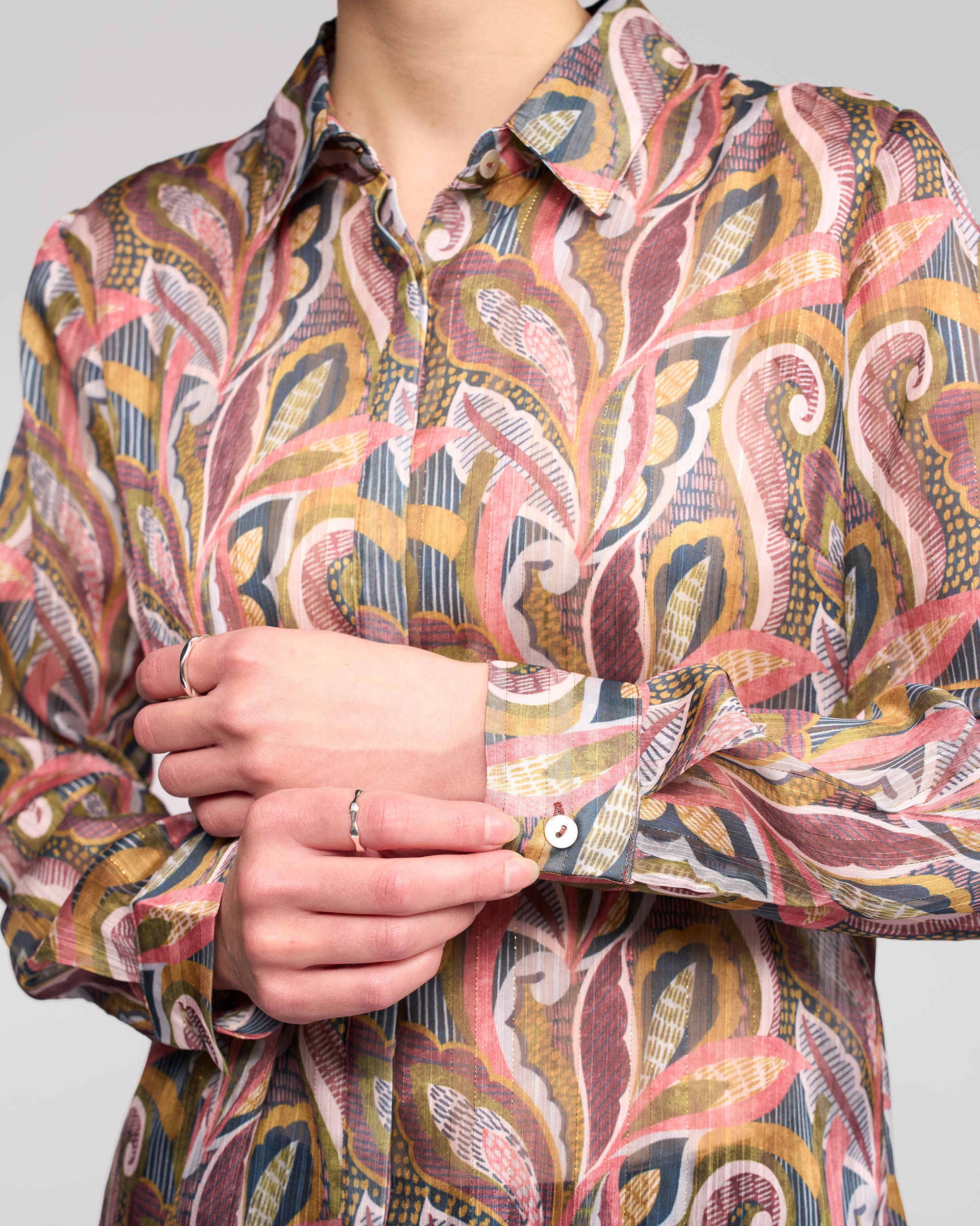 A person wearing the Vassalli Ballot Shirt - Sable, featuring long sleeves with pink, yellow, and green patterns, adjusts their cuff. Their torso and hands are visible, accessorized with several thin silver rings.